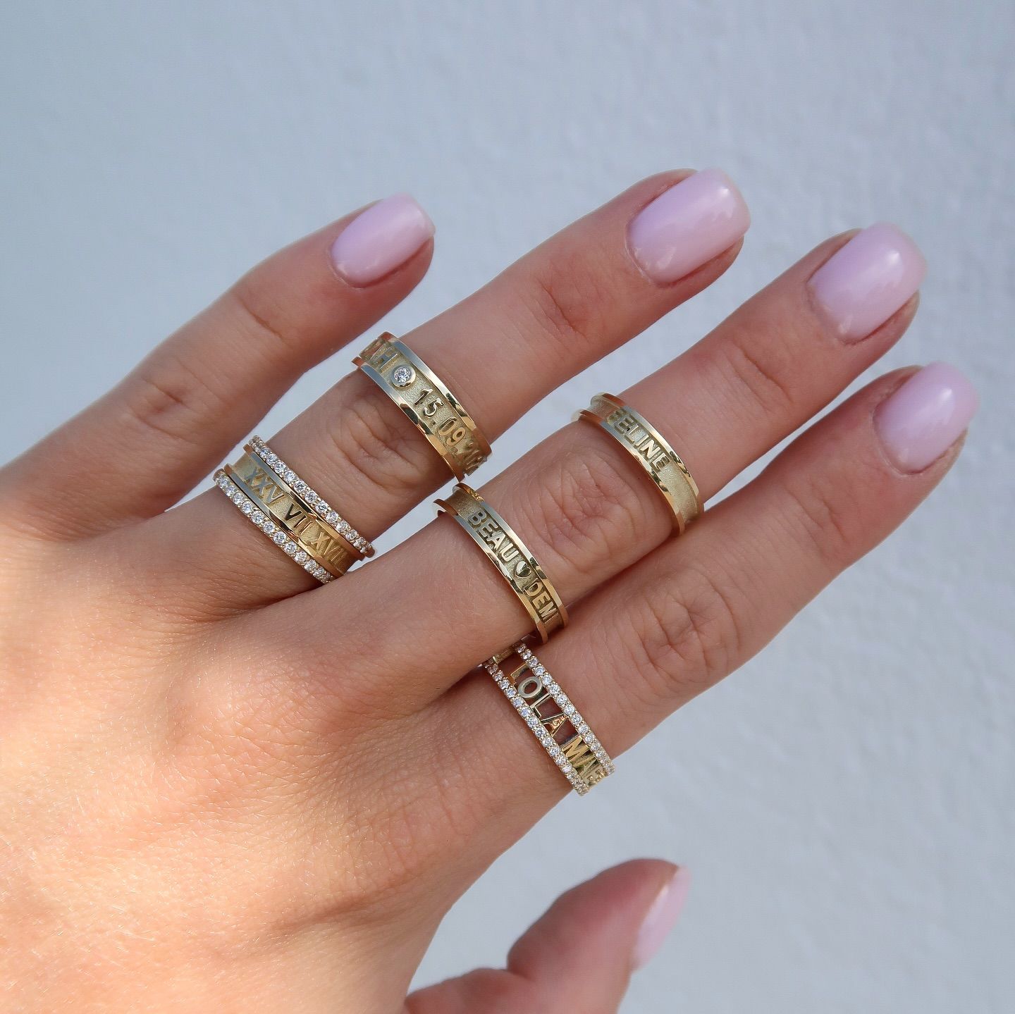 Hand met vijf gouden ringen, elk met een patroon, en lichtroze nagellak.