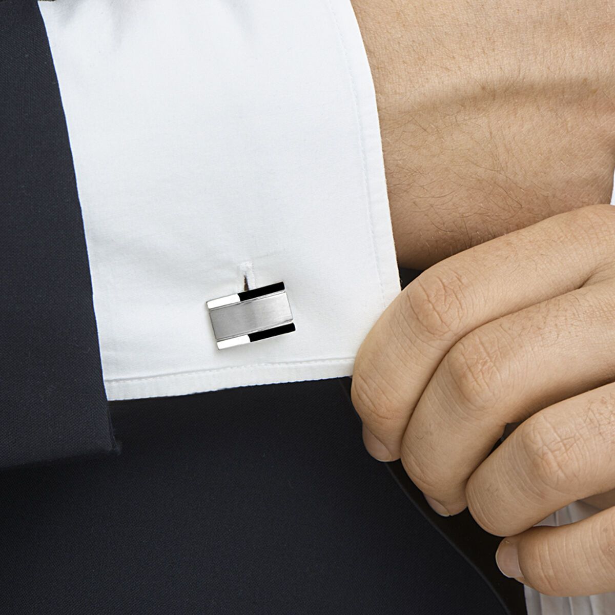 De hand van de man verstelt een manchetknoop van een overhemd; zilveren rechthoek met zwart accent; het colbert is zichtbaar.