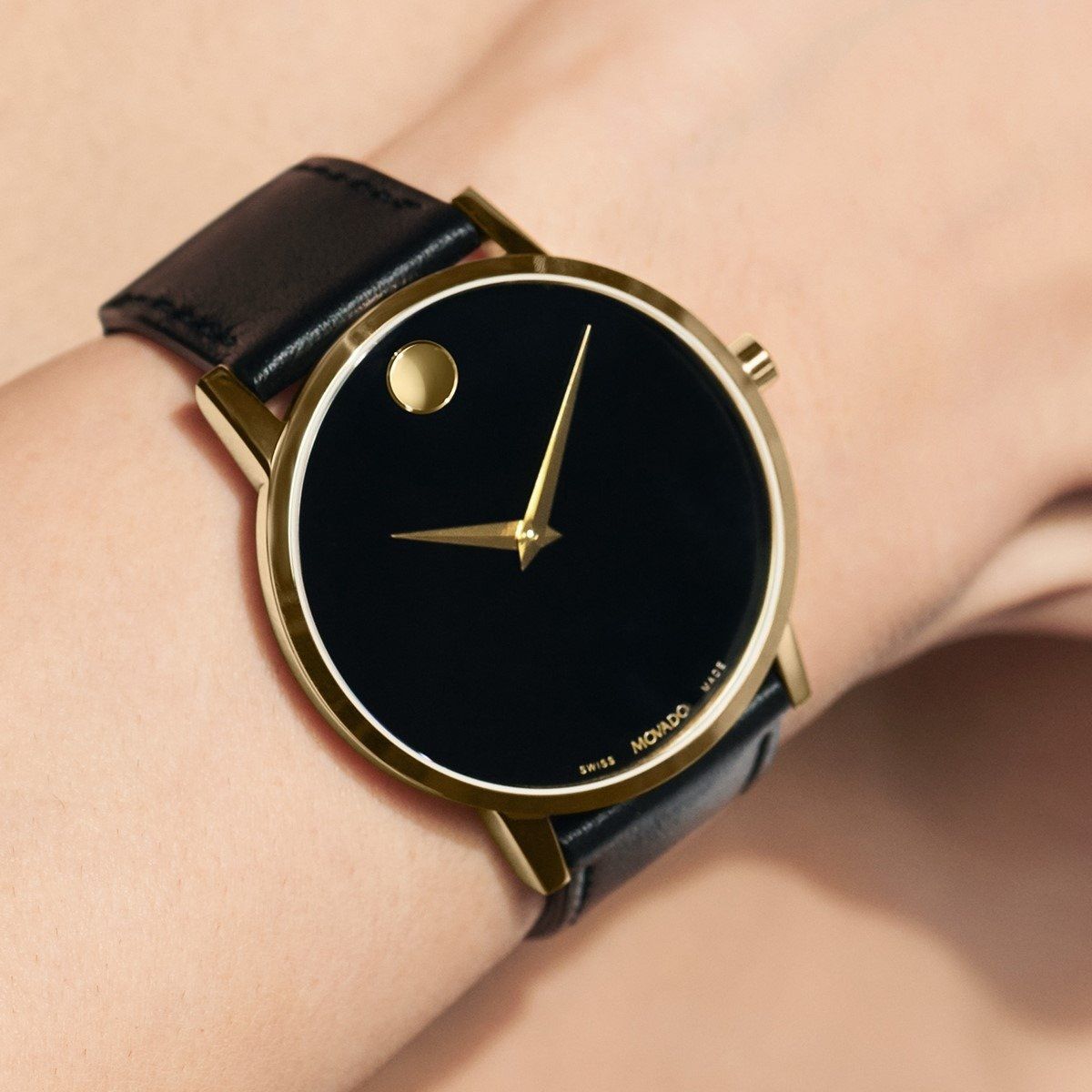 Movado horloge met gouden rand, zwarte wijzerplaat en gouden stip, gedragen om een pols met een zwarte leren band.