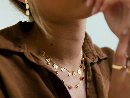Vrouw draagt ​​een gelaagde gouden ketting met rode edelstenen en bijpassende oorbellen, bruin topje.