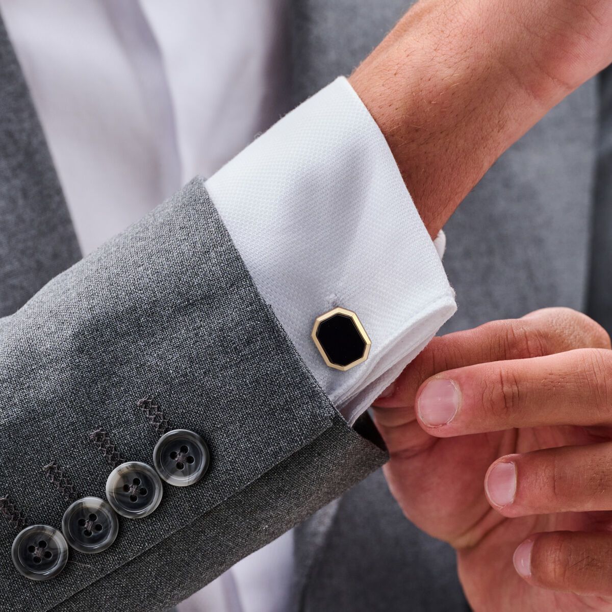 Man in grijs pak is bezig met het rechtzetten van een zwart-gouden manchetknoop op een wit overhemd.