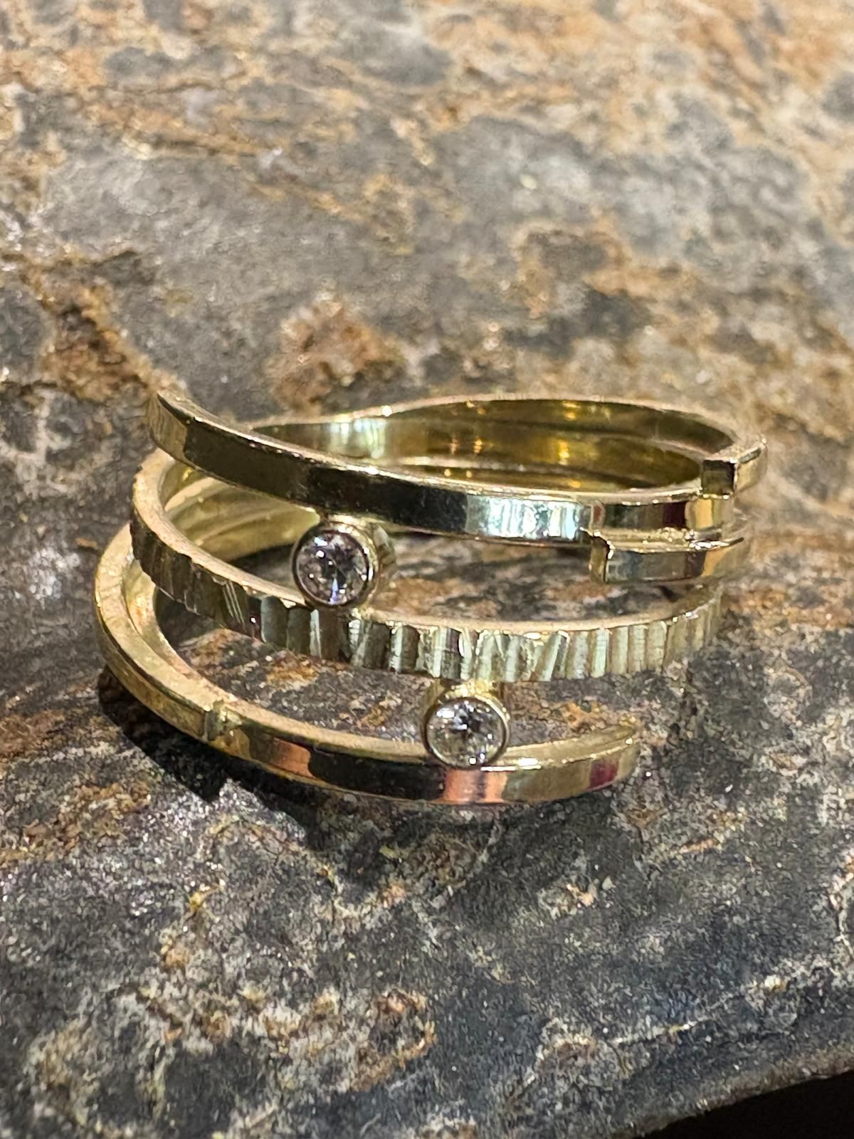 Gouden ring met drie overlappende banden, glad en gestructureerd, met twee kleine diamanten, rustend op een stenen oppervlak.