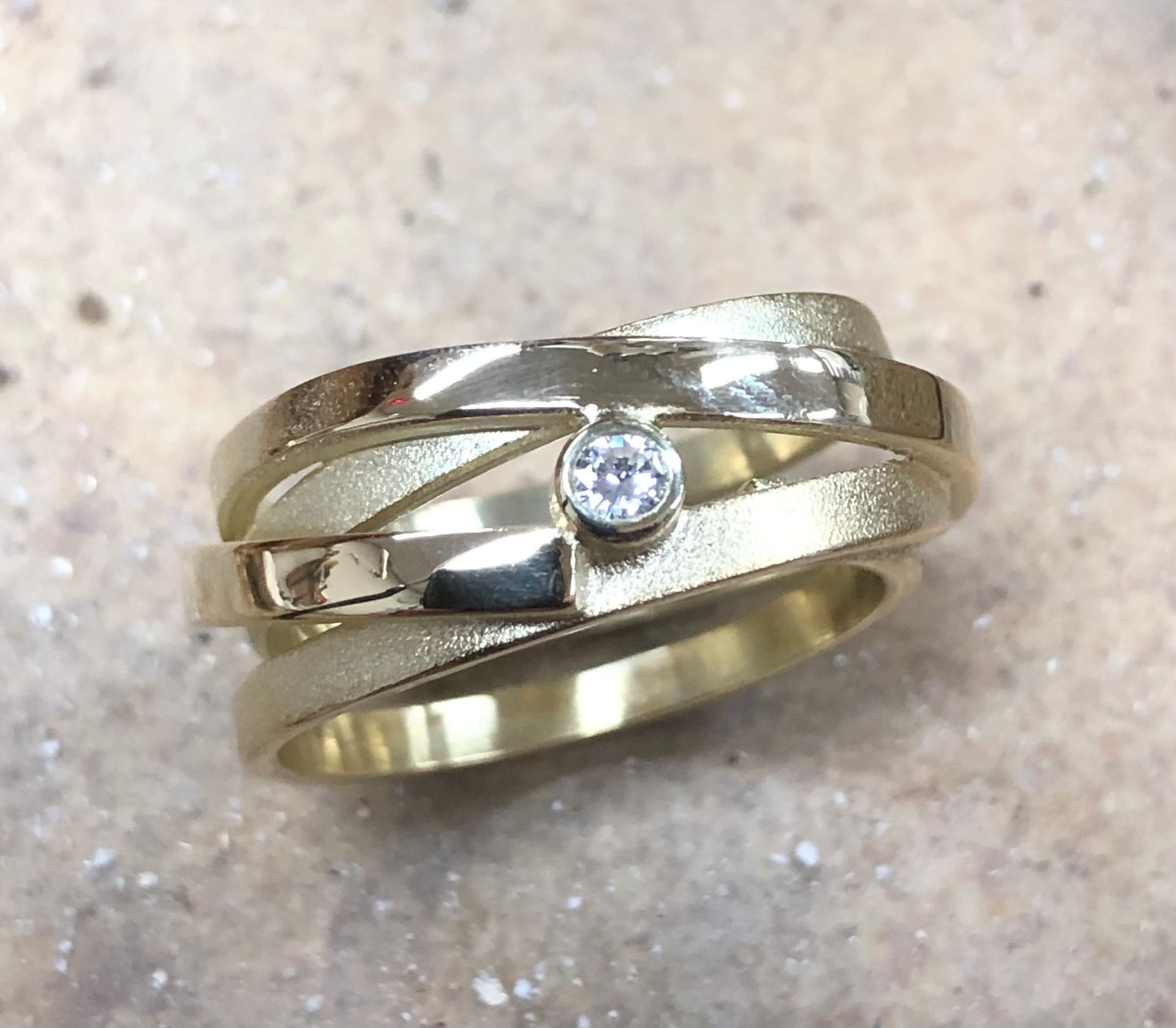 Goudkleurige ring met geweven design op een witte, vingervormige sieradenhouder tegen een donkergrijze achtergrond.