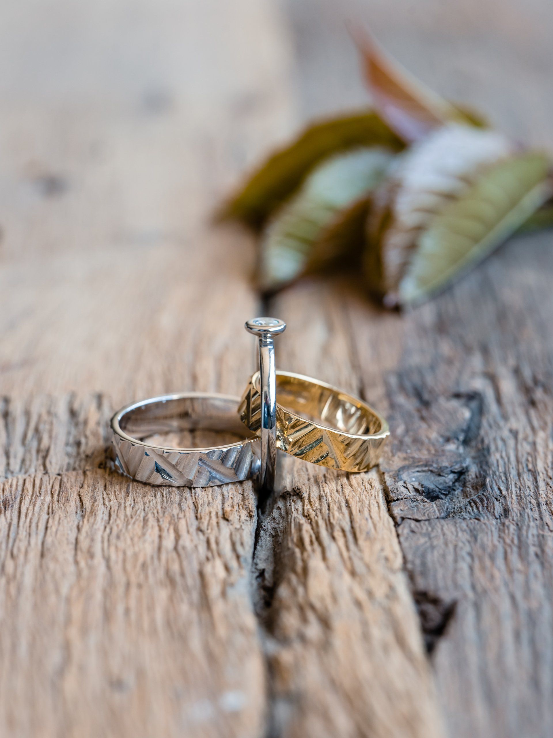 Trouwringen op verweerd hout met bladeren op de achtergrond. Een diamanten verlovingsring staat tussen twee getextureerde banden.