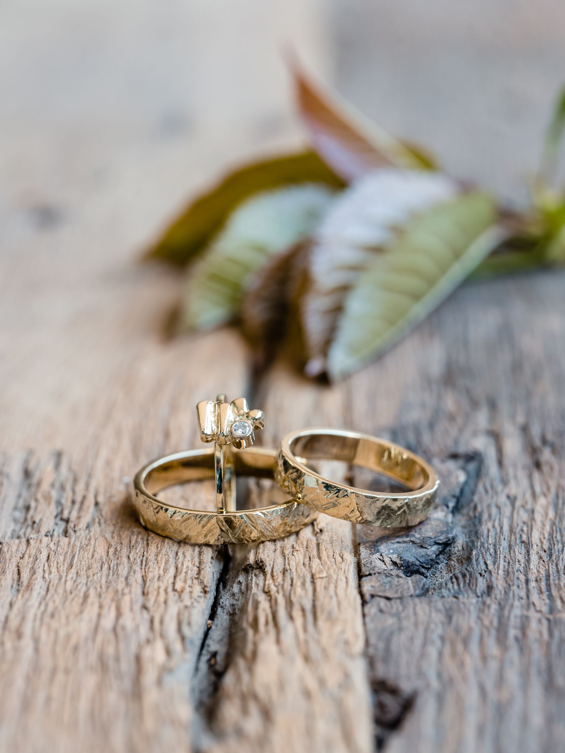 Drie getextureerde gouden trouwringen op een verweerd houten oppervlak, met een bladtakje op de achtergrond.