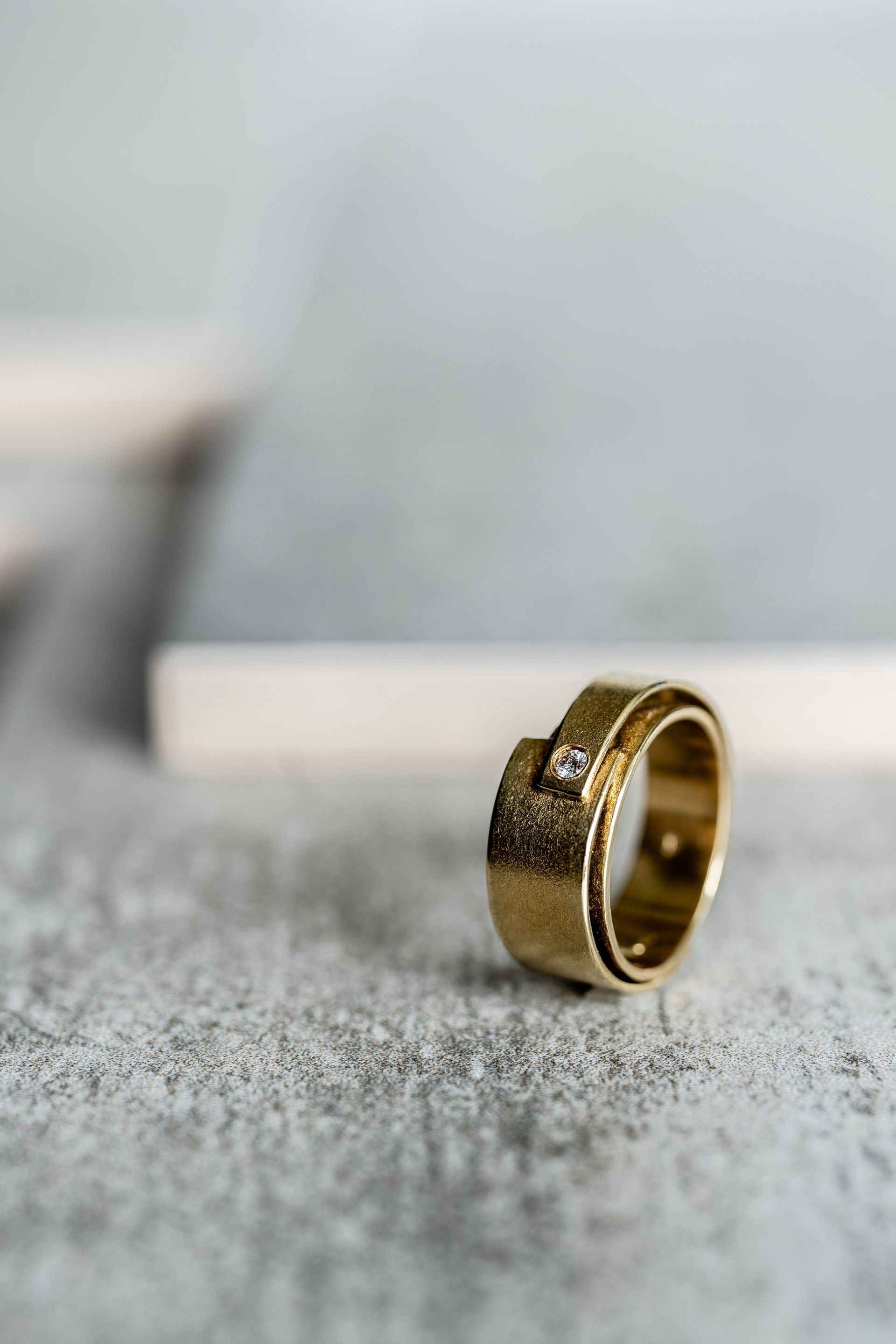 Gouden ring met structuur en een kleine vierkante edelsteen, rustend op een grijs oppervlak.