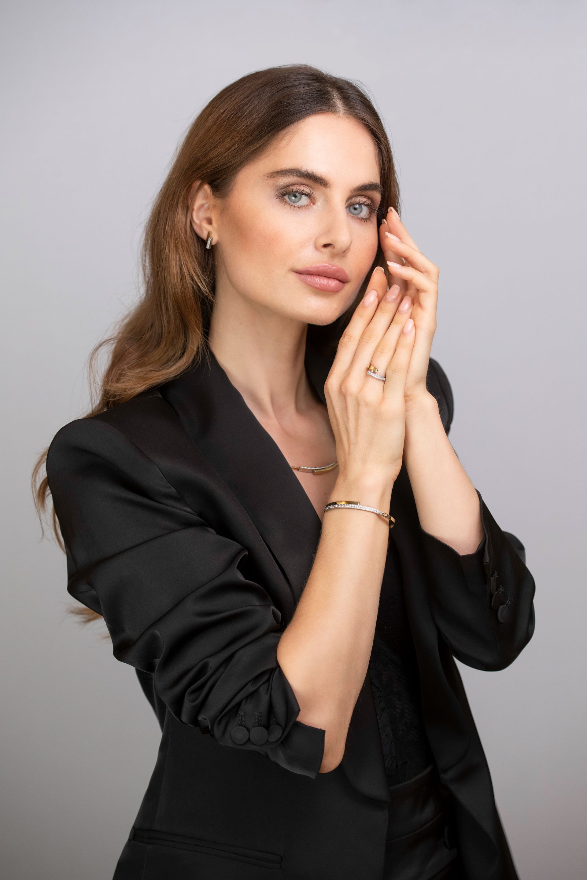 Vrouw in een zwart jasje, met sieraden, poserend met haar handen dicht bij haar gezicht.