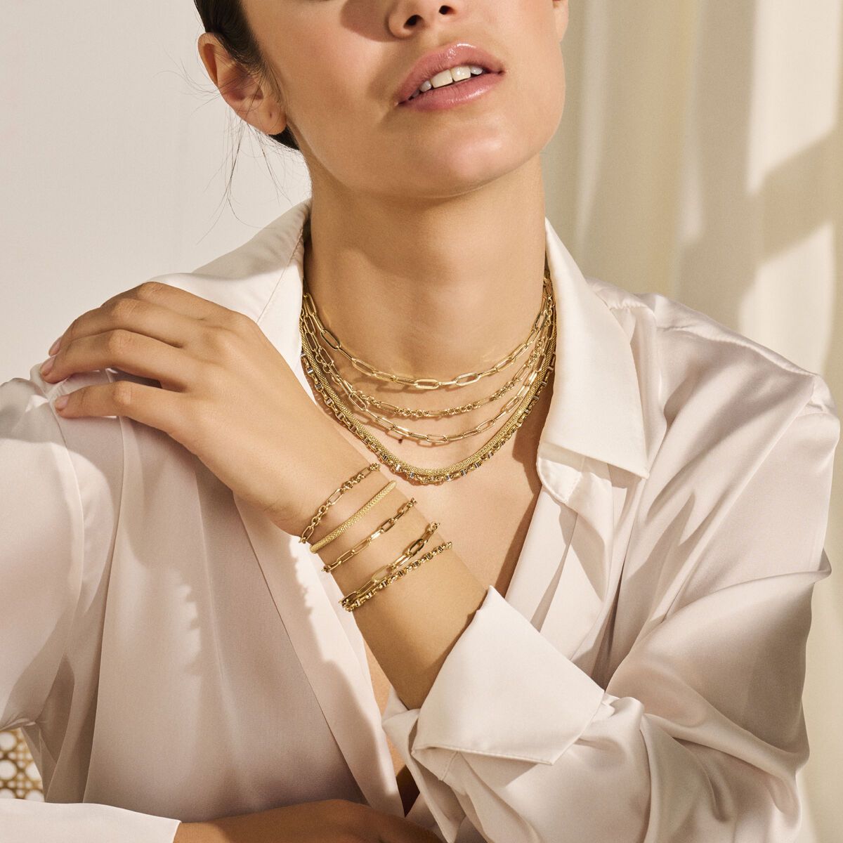 Vrouw draagt ​​meerdere gouden kettingen en armbanden en een wit overhemd.