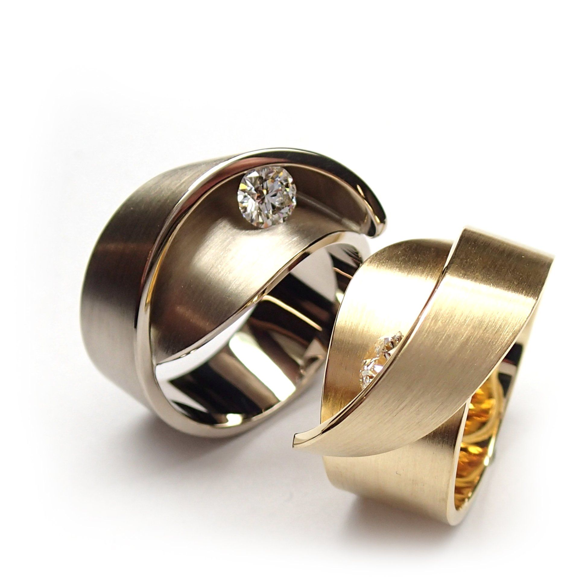 Twee trouwringen, een zilveren met een diamant, een gouden met een diamant. Beide hebben een wervelend, lintachtig ontwerp.