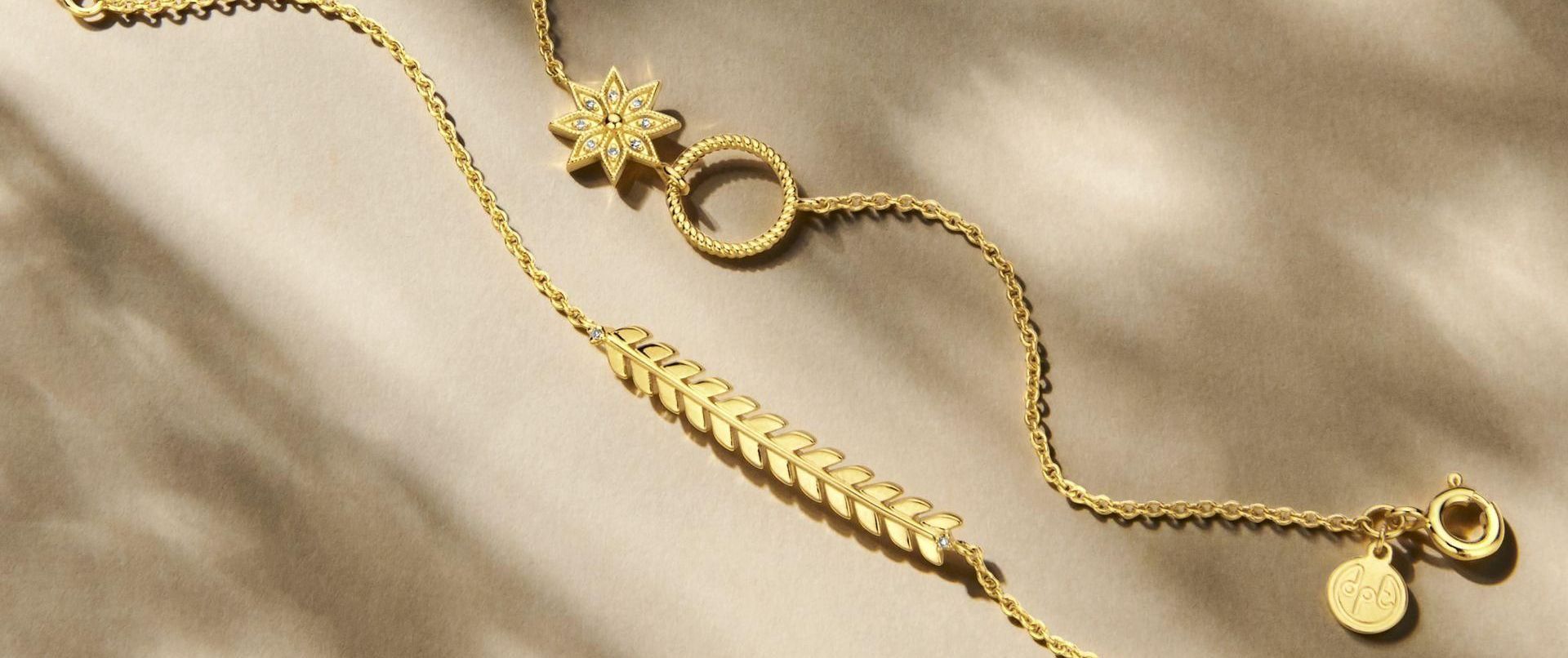 Gouden armbanden op een gestructureerde achtergrond. De ene heeft een bloemenbedeltje, de andere een bladmotief.