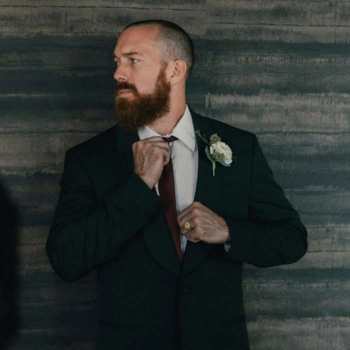 Man in donker pak, met donkere baard en wit corsage, doet zijn stropdas goed.