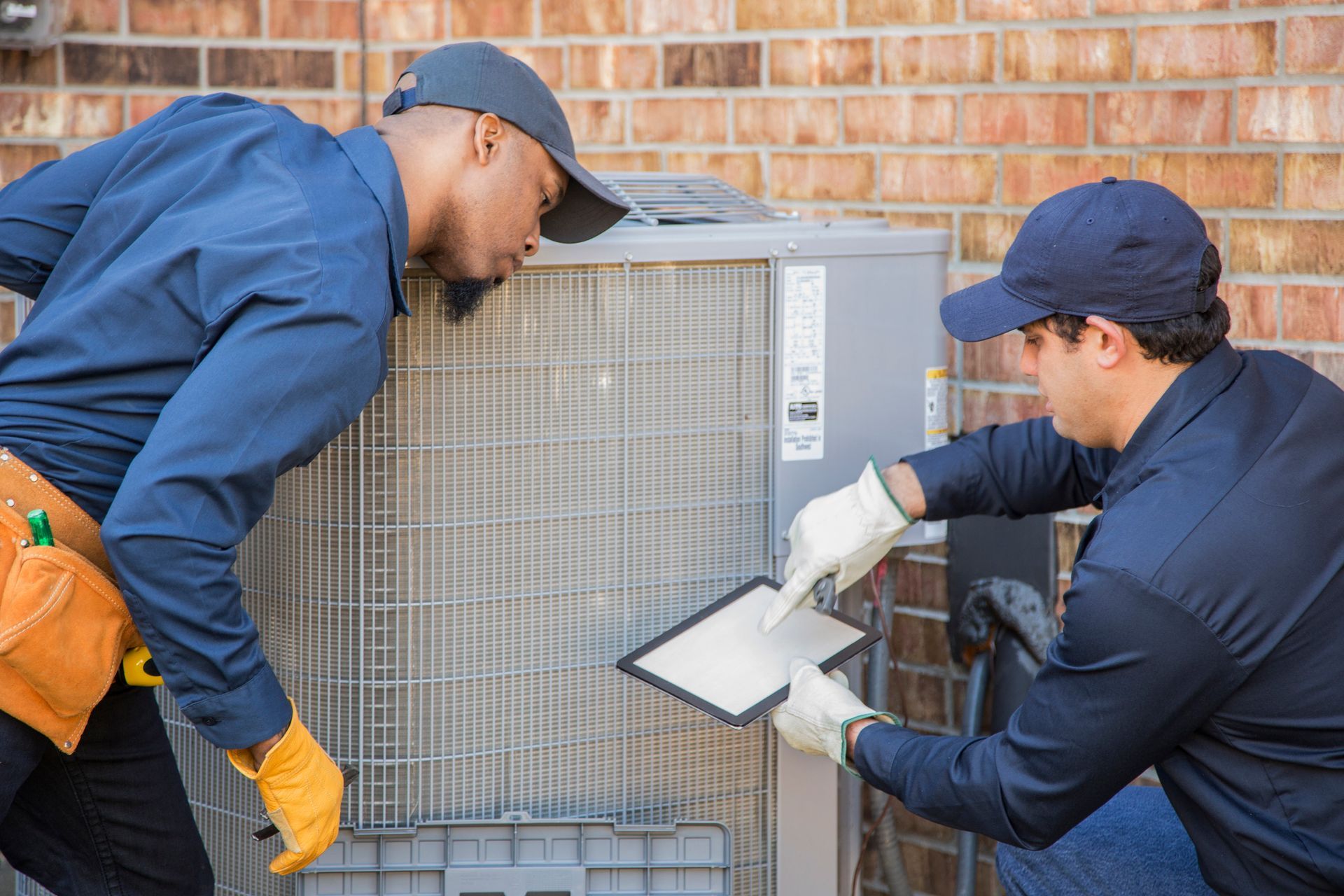 Two technicians in blue uniforms inspecting an outdoor AC unit against a brick wall. One holds a tablet.