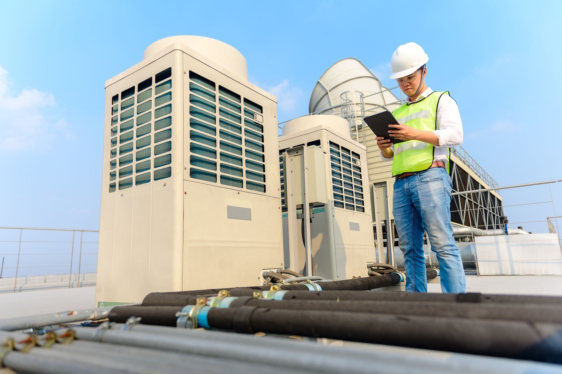 An engineer in a hard hat and vest examines rooftop HVAC units using a tablet.