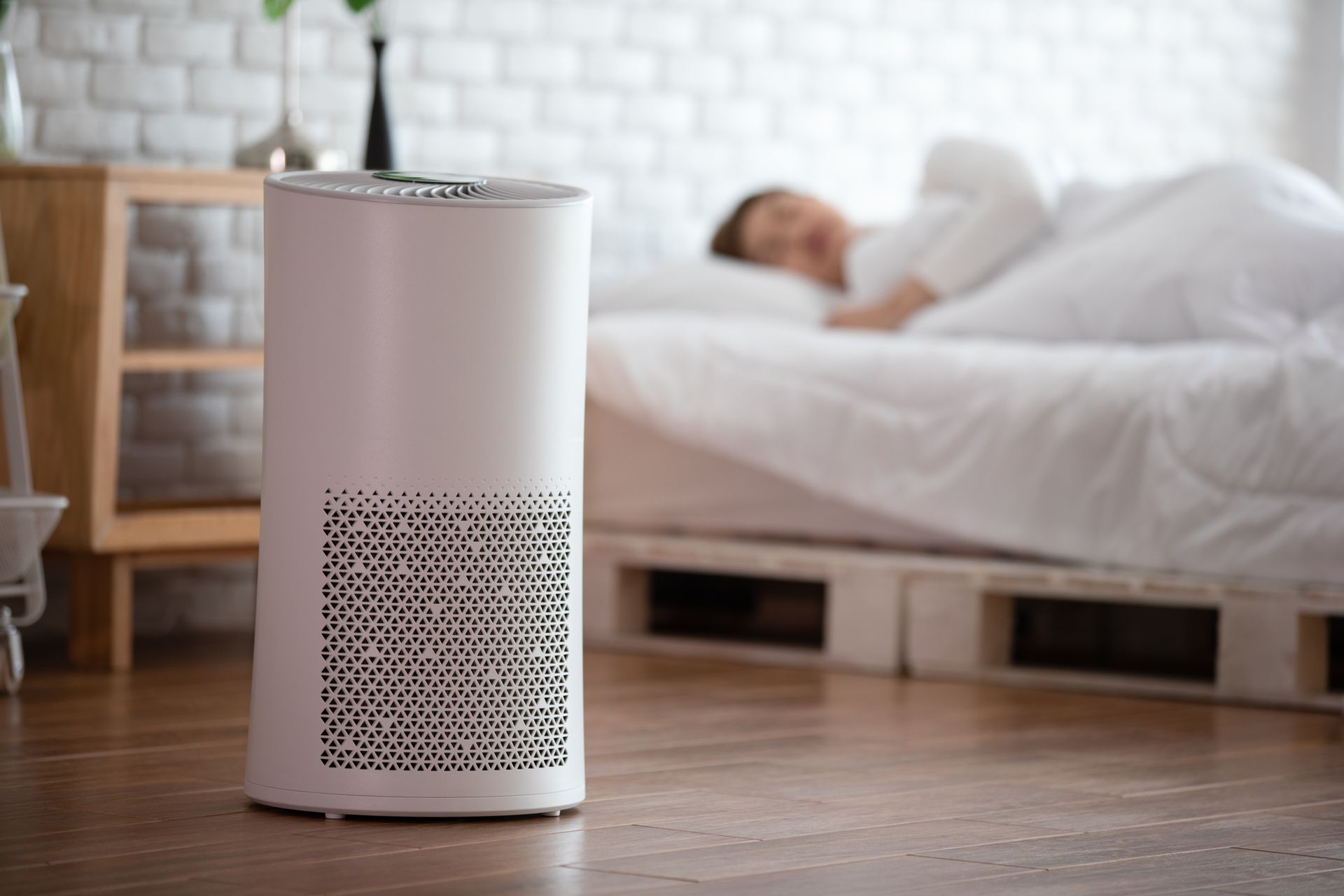 Air purifier in a bedroom; person sleeping in bed. White machine near wooden nightstand and person.