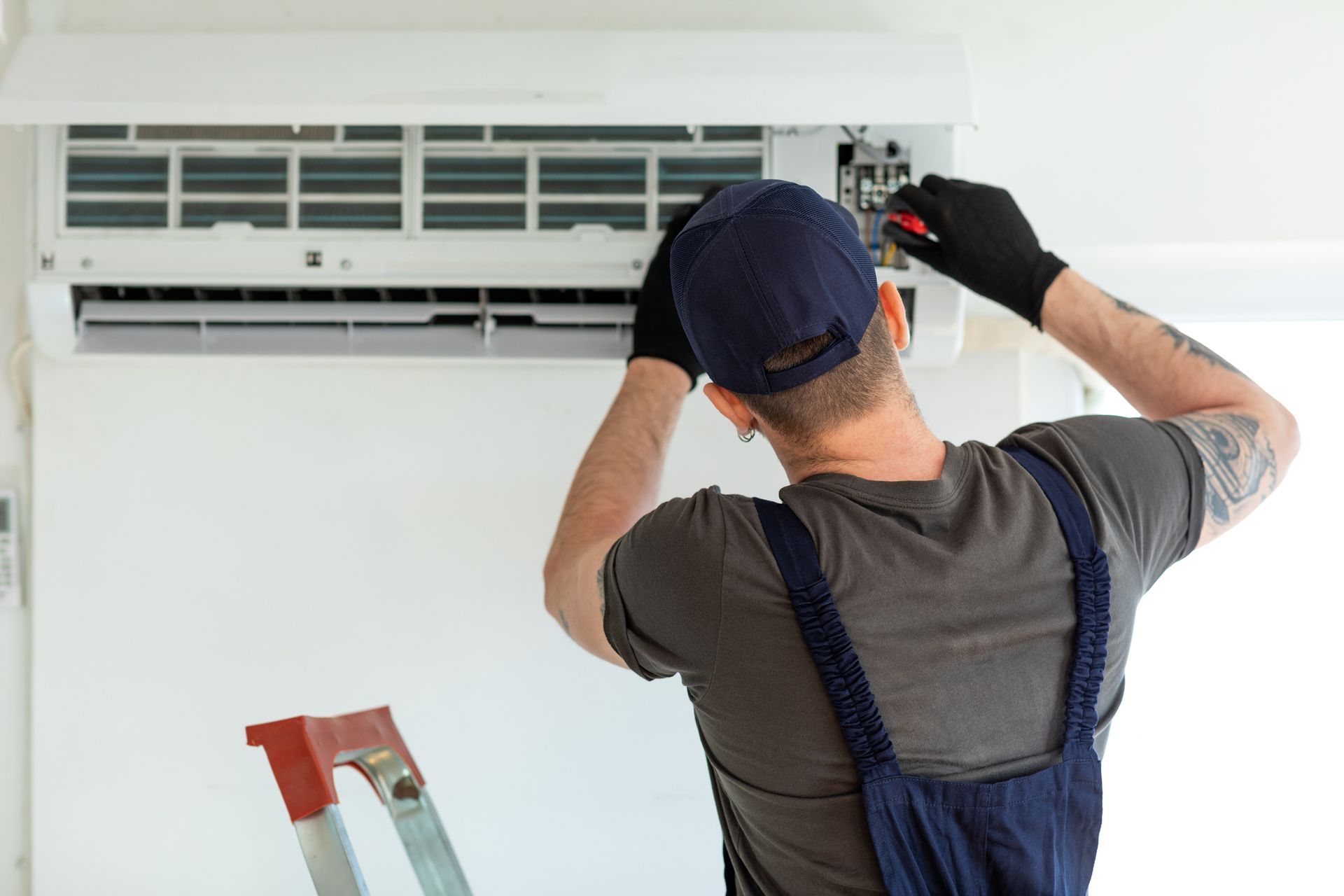 A technician in a cap and overalls repairs a wall-mounted AC unit, using tools.