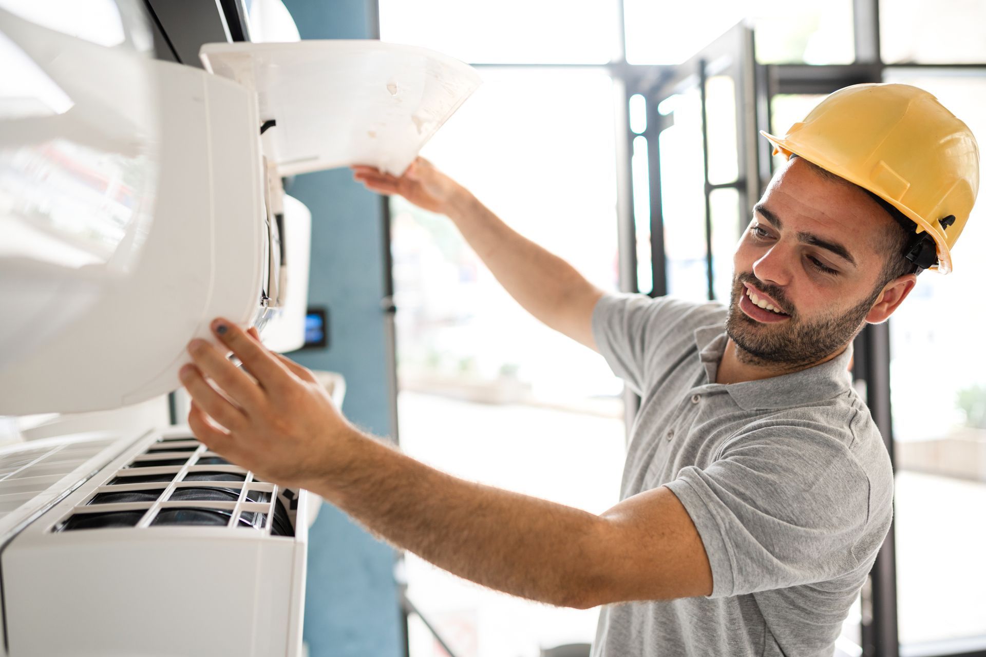 Man in hard hat servicing an air conditioner; smiling, working indoors.