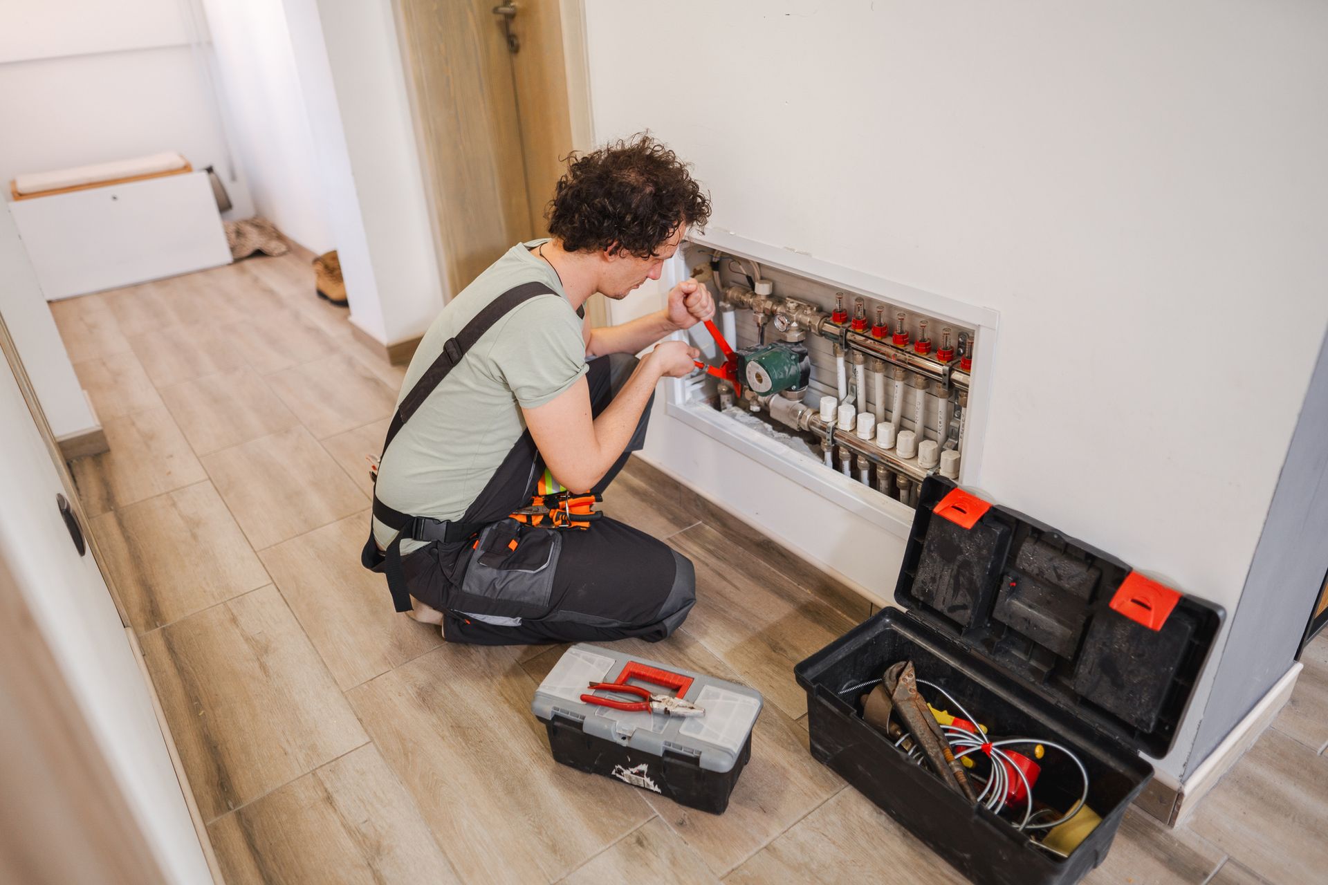 Person in work overalls repairing plumbing fixtures in a wall niche, tools visible.