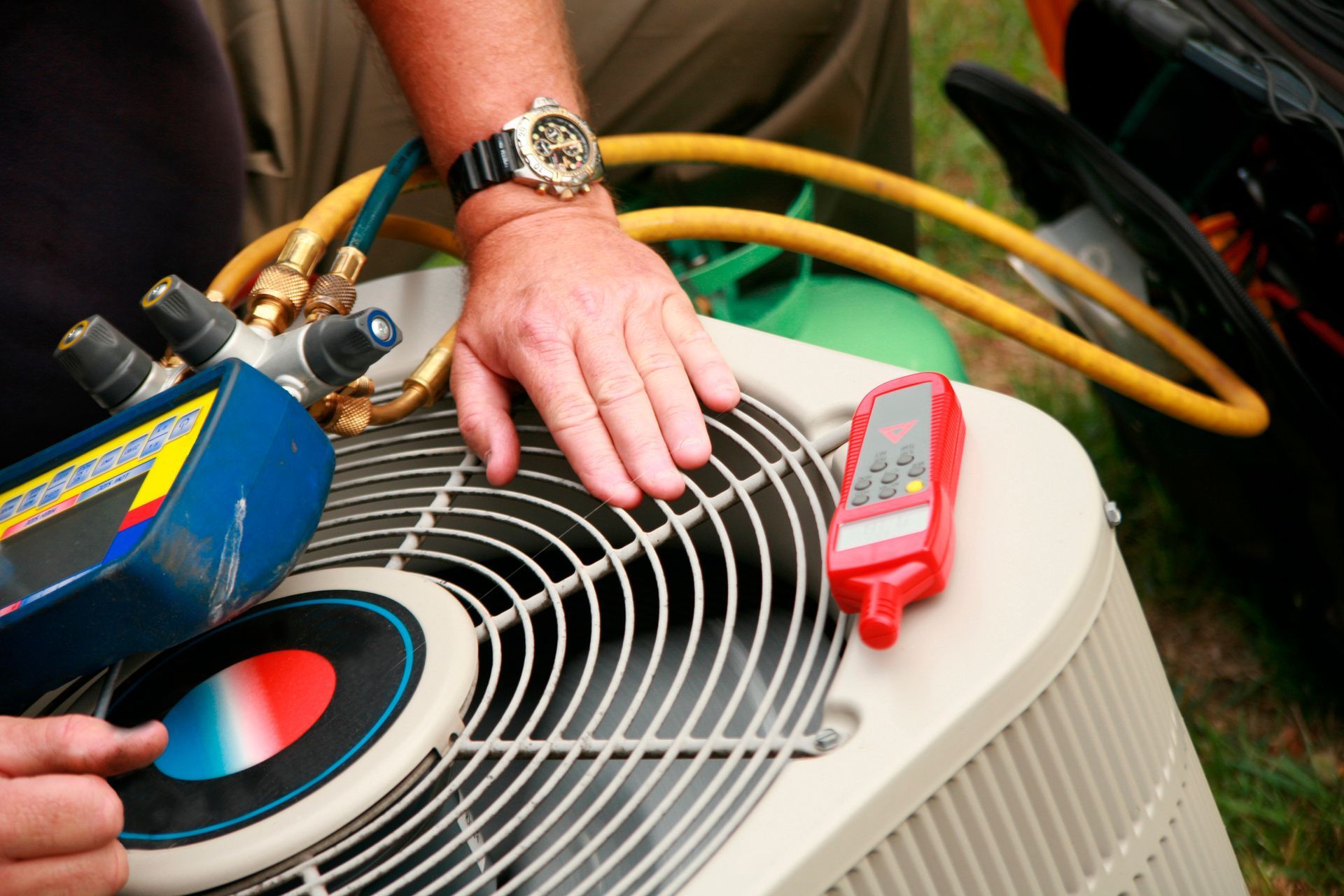 Person's hand on AC unit with gauges and tools, connected by yellow hose.  A green tank is in the background.