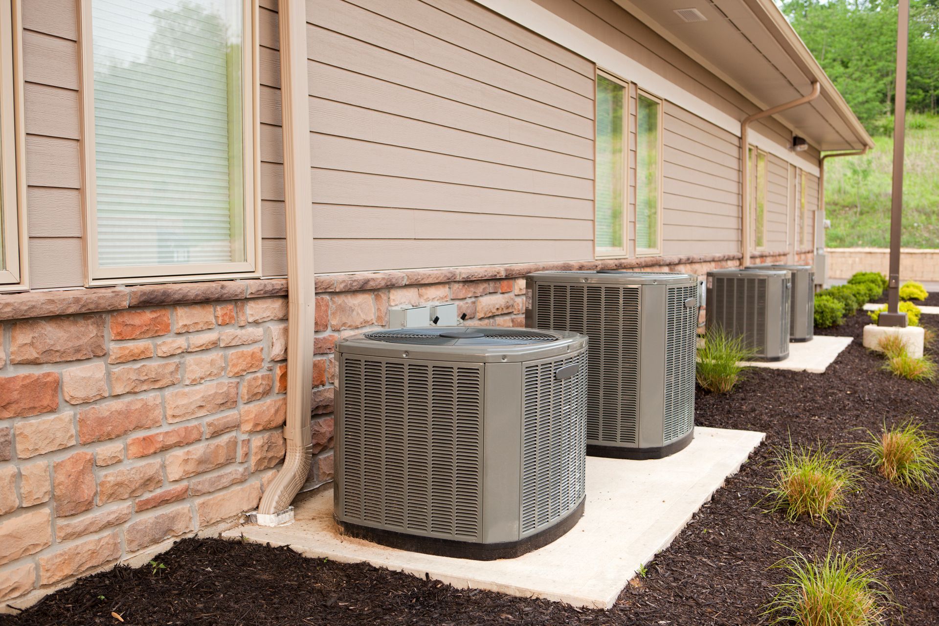 Air conditioning units lined up outside a building with brick and beige siding.