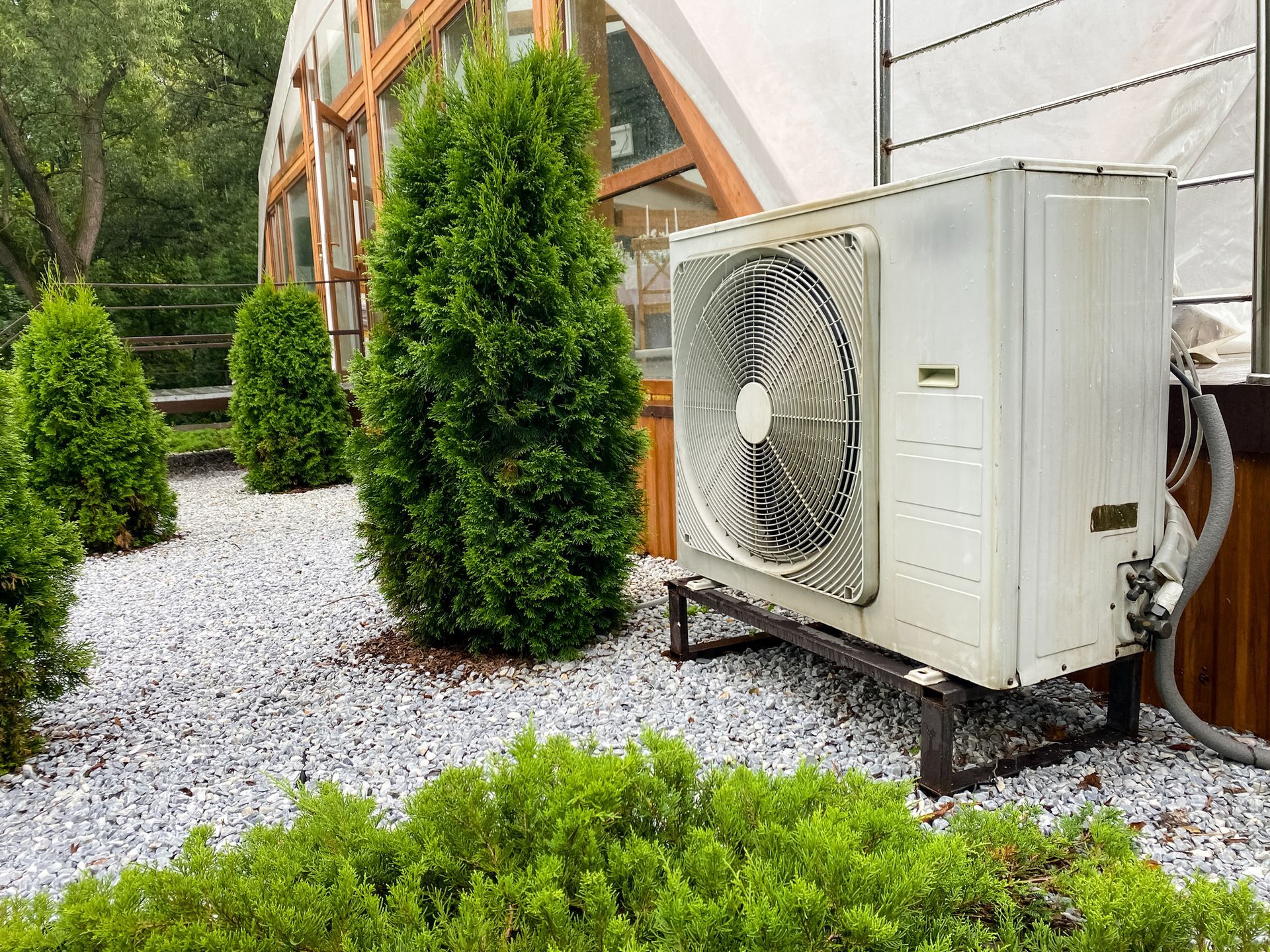 HVAC unit outside of a building on a metal stand surrounded by landscaping with small trees and white gravel.
