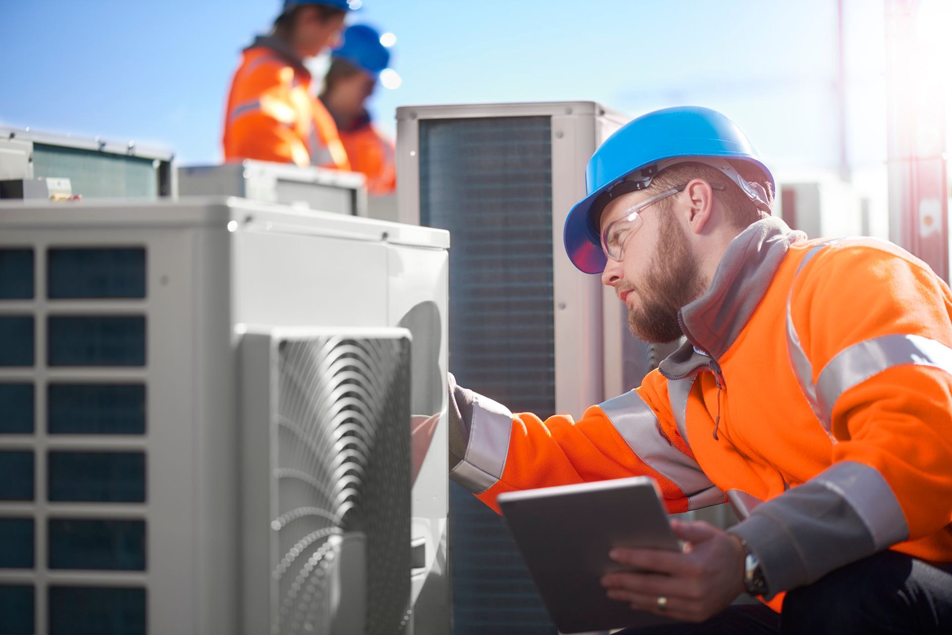 HVAC technician in orange safety gear inspecting an air conditioning unit, using a tablet, outdoors.