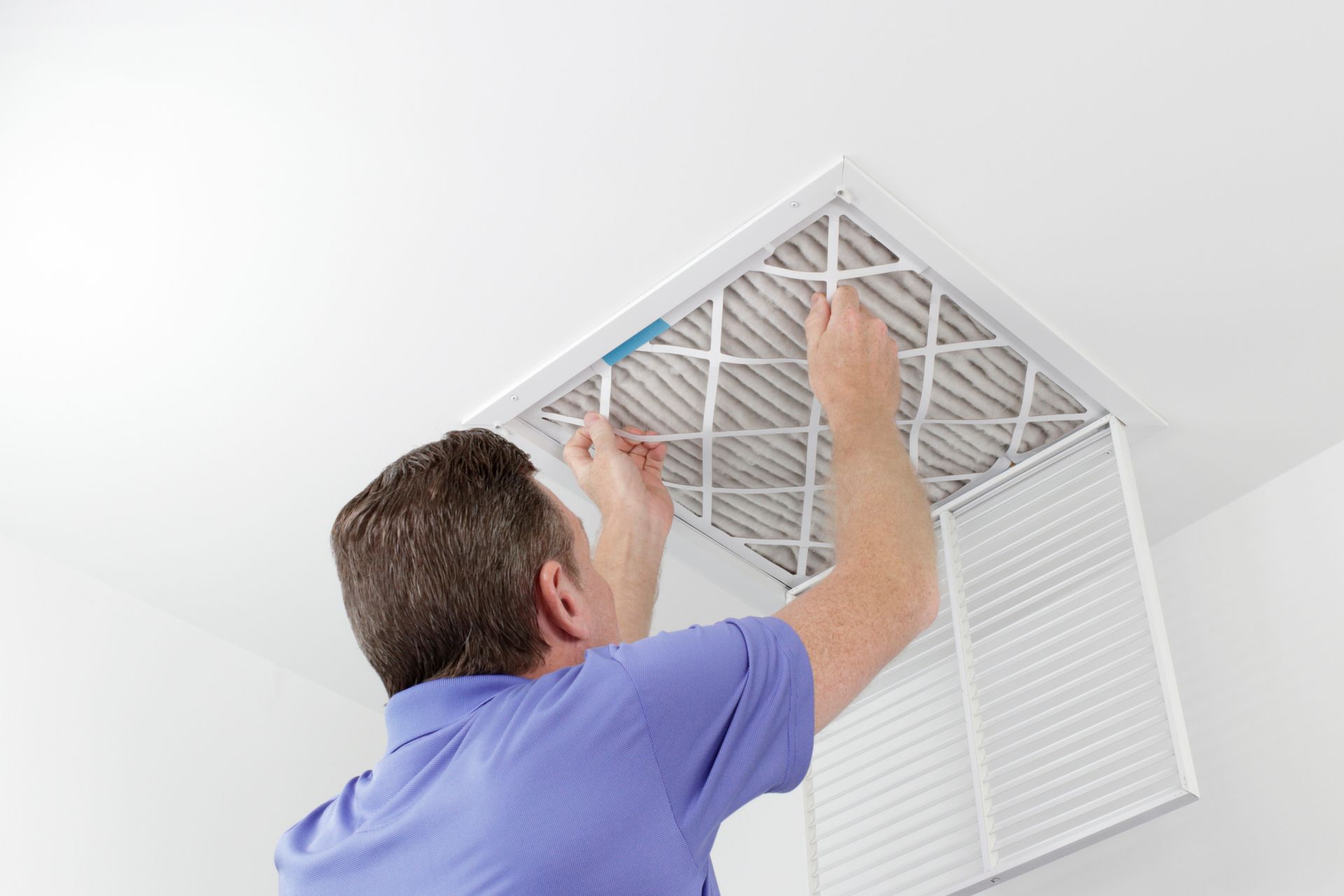 Man replacing an air filter in a ceiling vent; white room, light blue shirt.