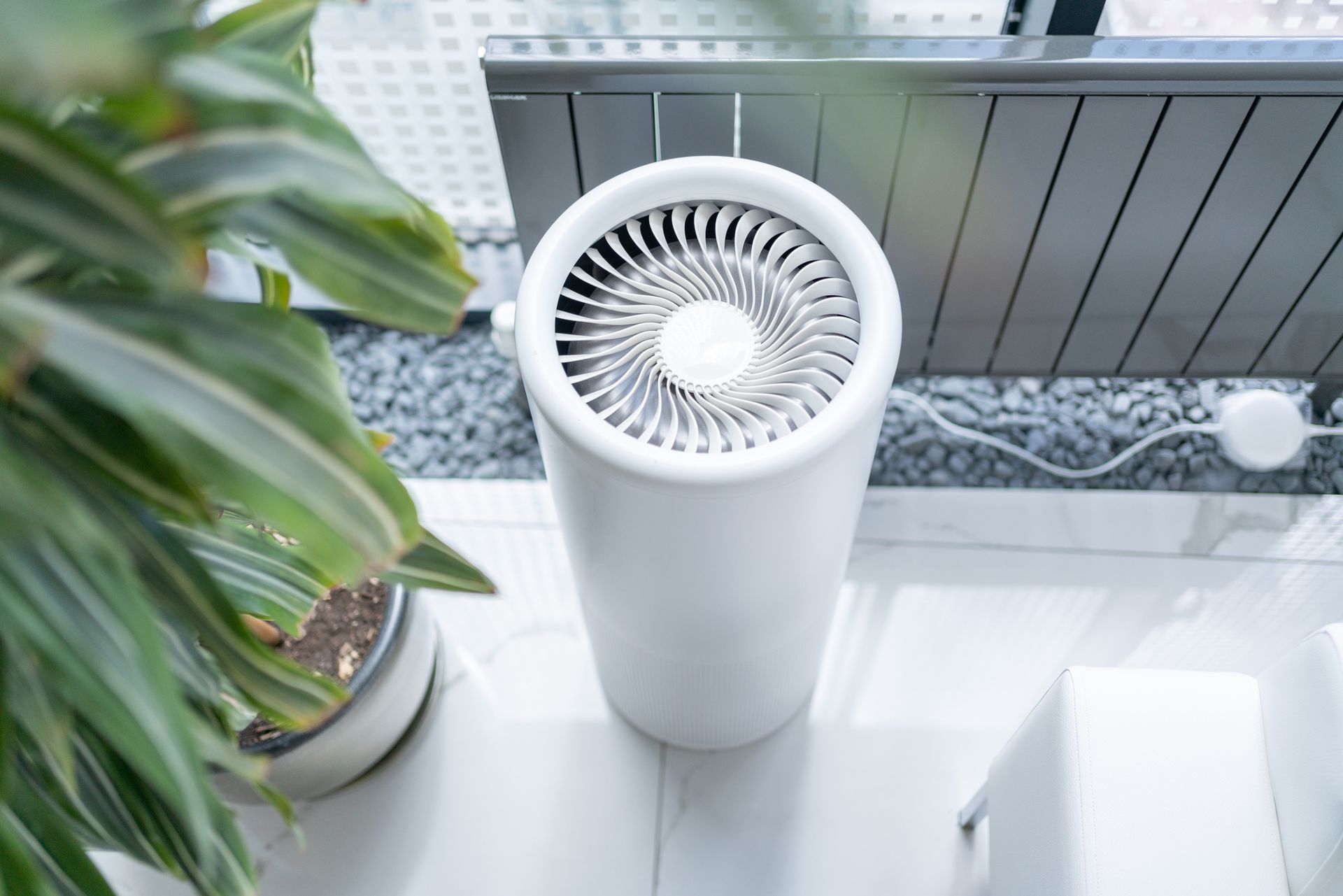 White air purifier on white tile floor next to a green plant, with a window and outdoor seating in the background.
