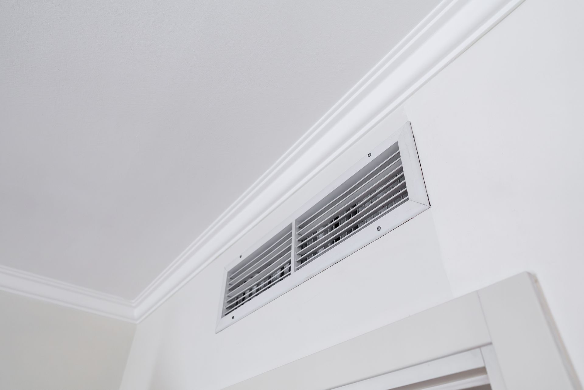 White rectangular air vent on a white wall below a white ceiling with crown molding.