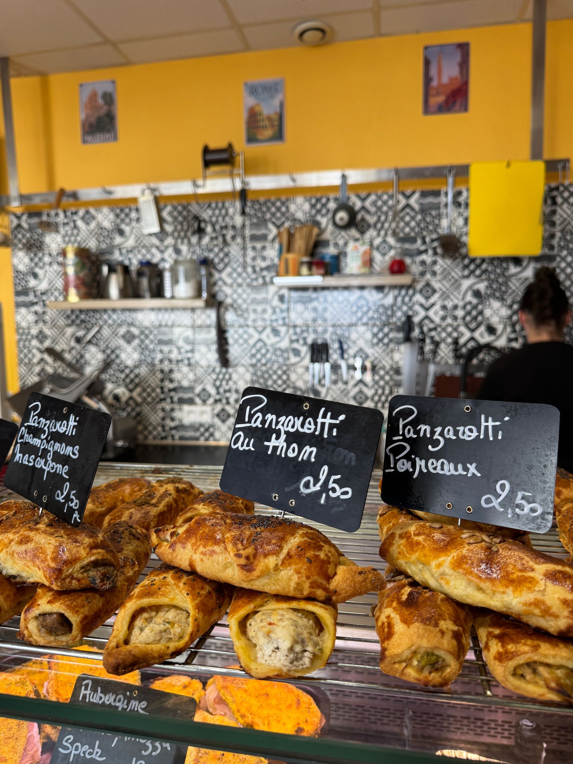 Des pâtisseries étiquetées sont exposées dans une boulangerie. Une personne travaille derrière le comptoir. Mur jaune et carrelage à motifs.