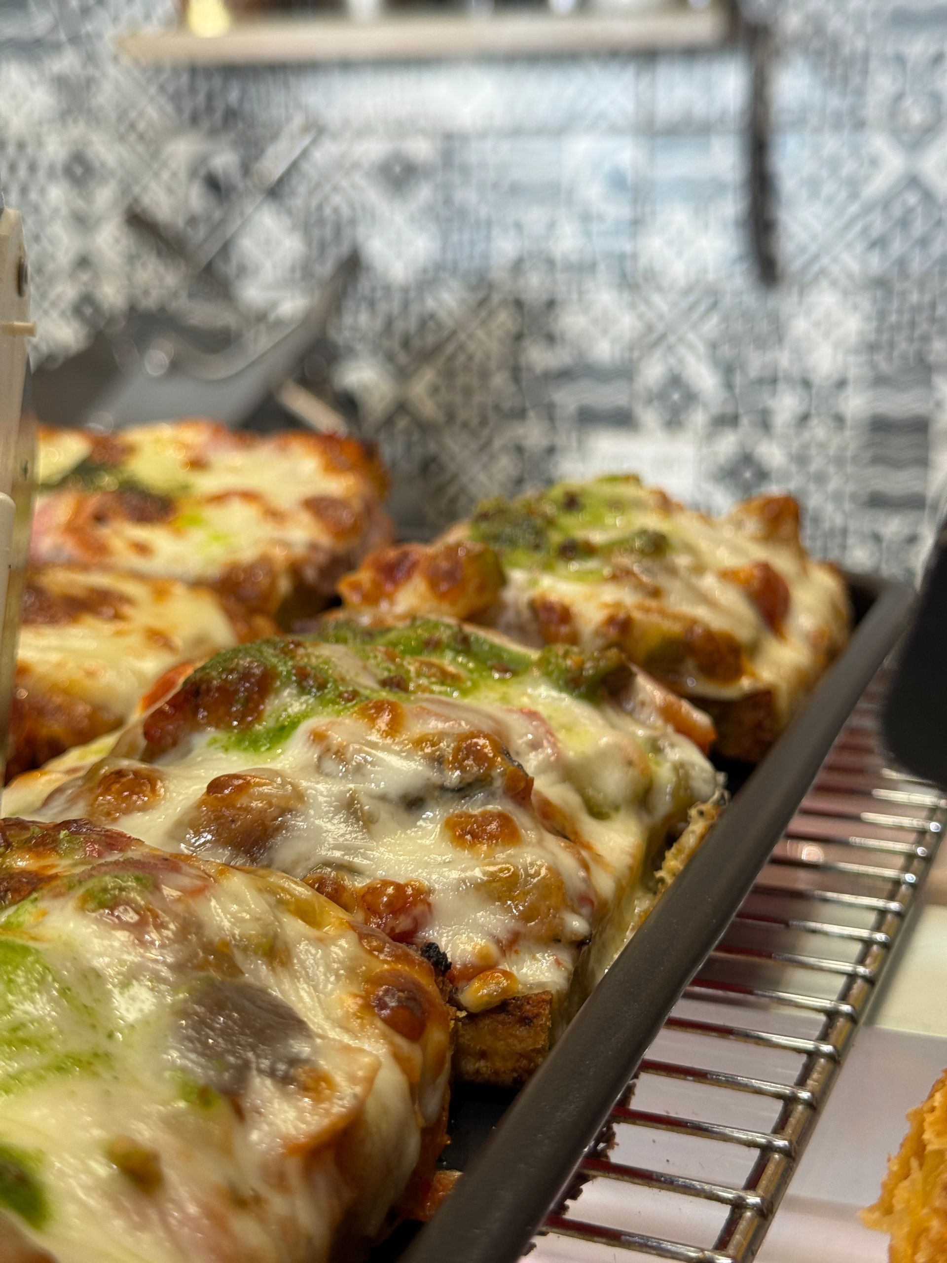 Des tartines avec du fromage fondu et des garnitures, présentées sur un support métallique, avec un mur à motifs en arrière-plan.