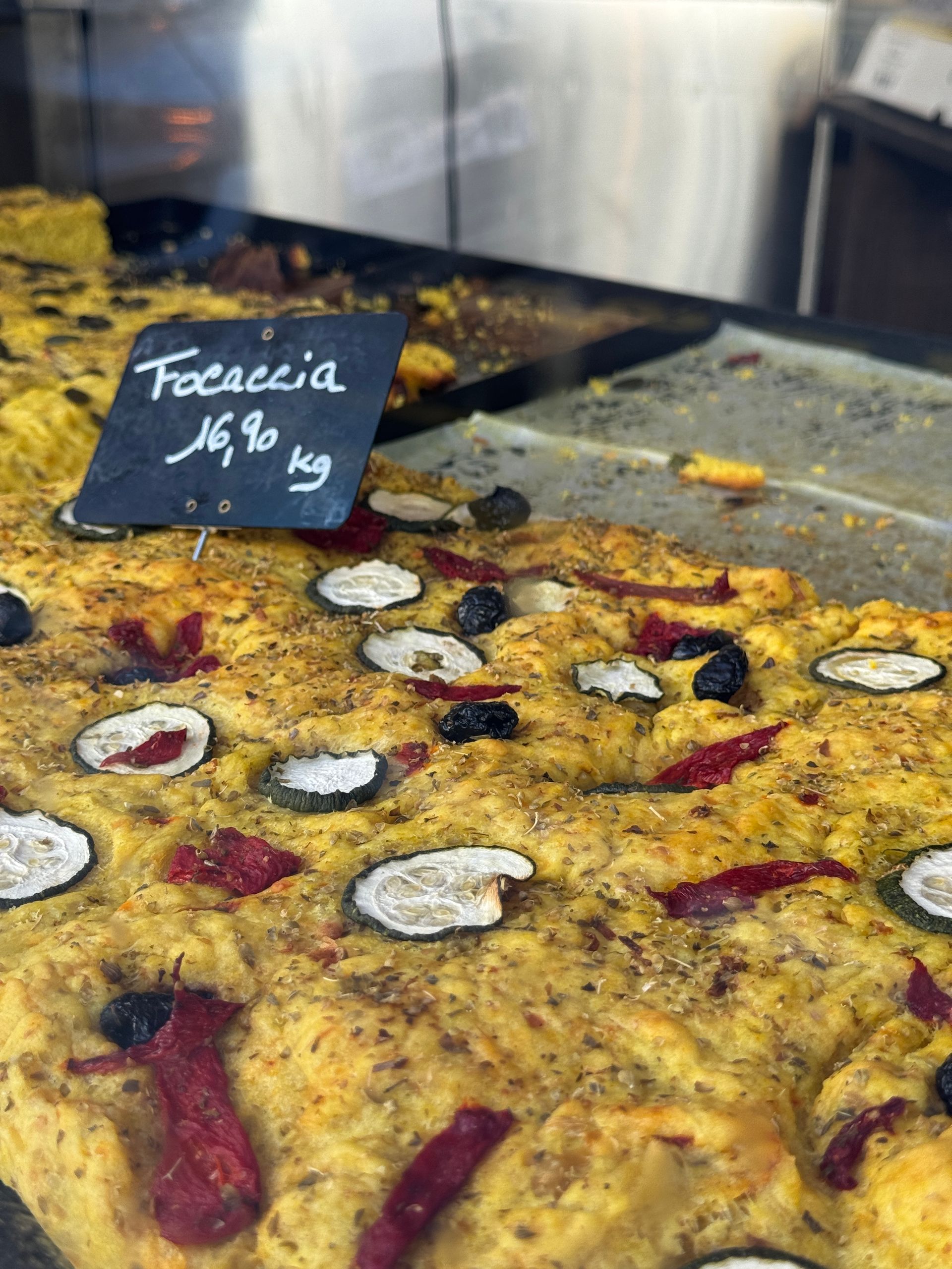 Pain focaccia garni, dans une vitrine ; une pancarte indique « Focaccia 16,90 kg ».
