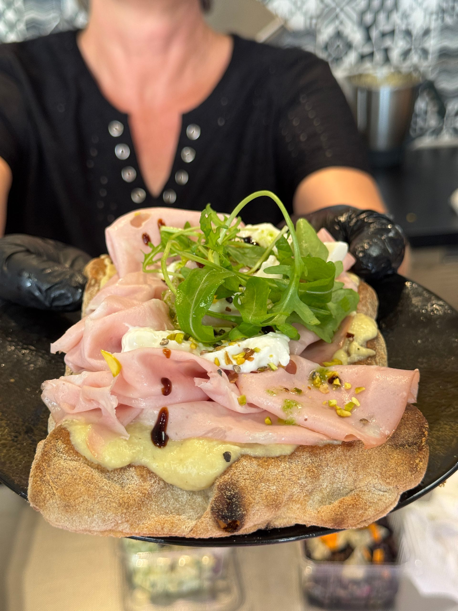 Sandwich servi sur une assiette noire par des mains gantées, garni de viande, de fromage, de verdure et de pistaches.