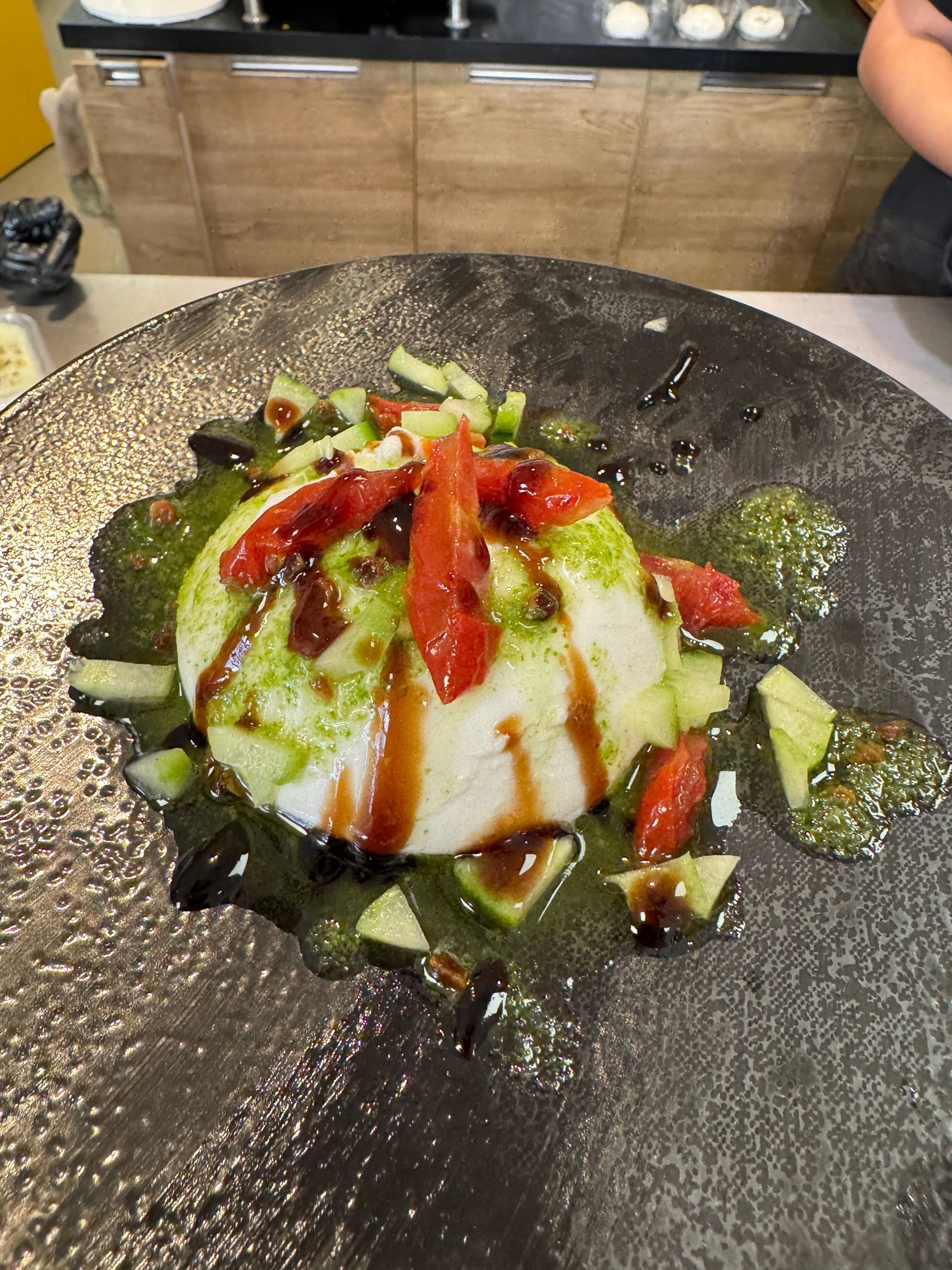 Burrata avec tomates hachées, concombre, pesto et réduction balsamique sur une assiette foncée.