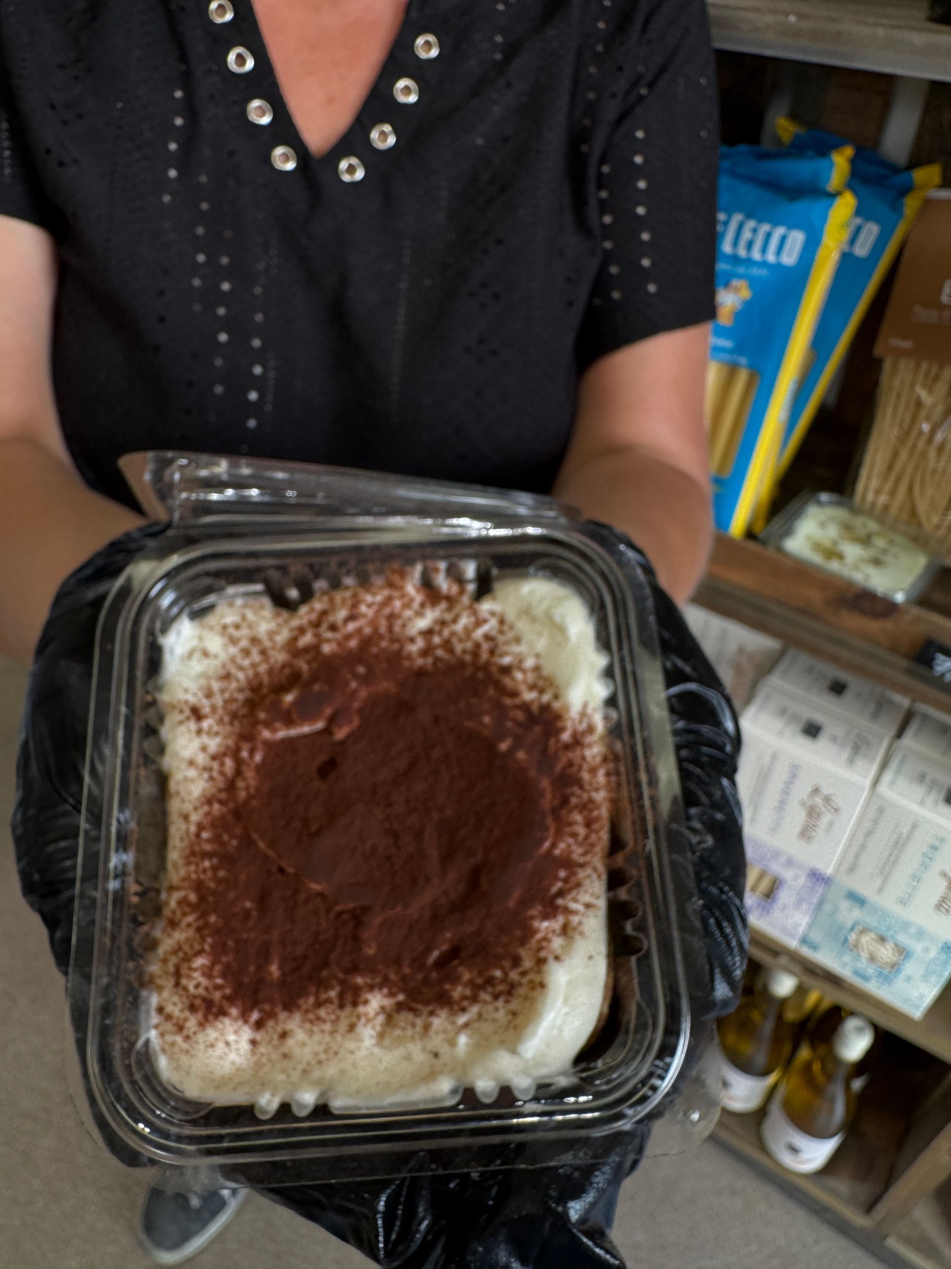 Une personne tenant un tiramisu dans un récipient en plastique transparent, saupoudré de cacao en poudre.