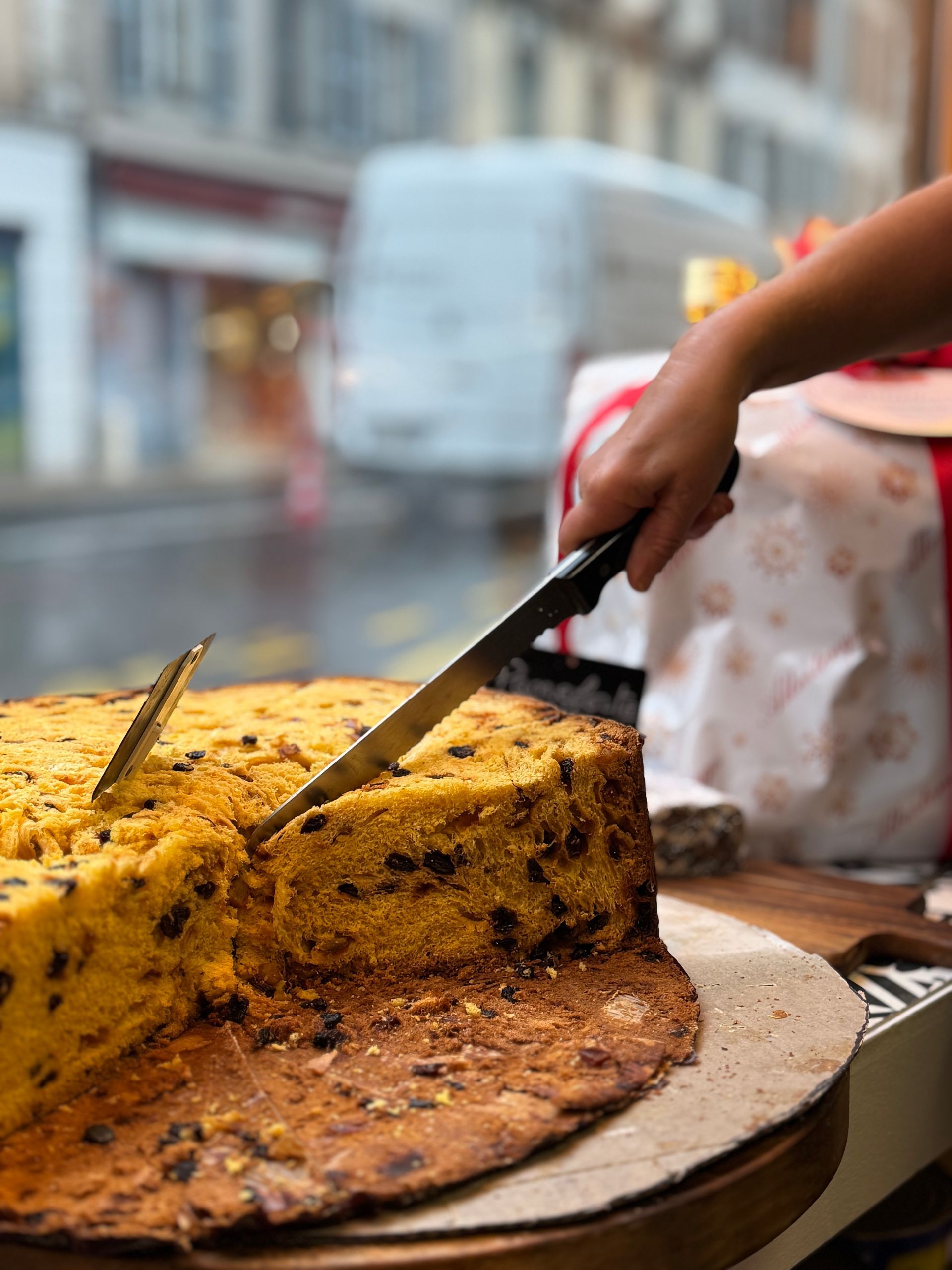 Une main découpant un gâteau rond et jaune parsemé de morceaux foncés, dans la vitrine d'un magasin.