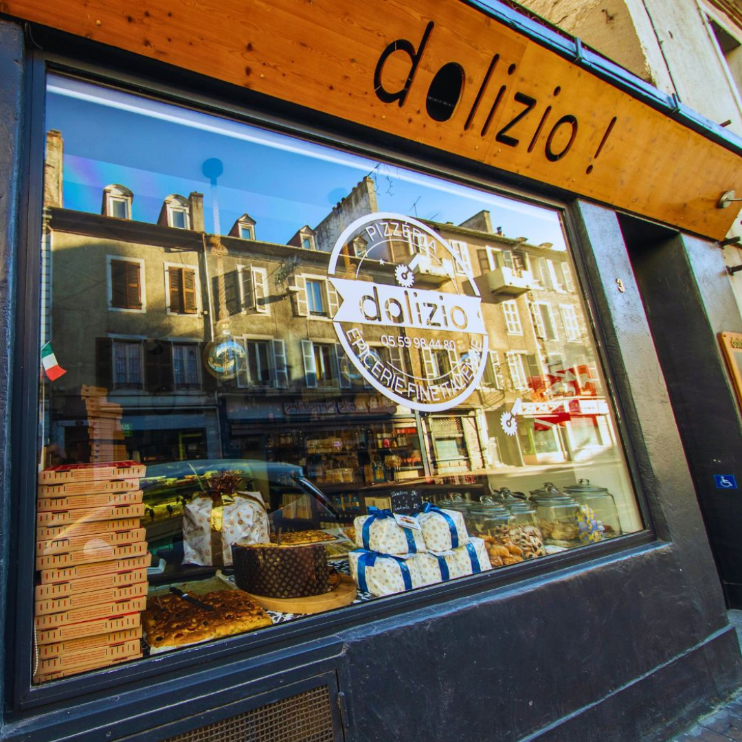 Extérieur d'une épicerie fine italienne « dolizio ! » avec une vitrine présentant des produits alimentaires ; reflets de bâtiments dans la vitrine.