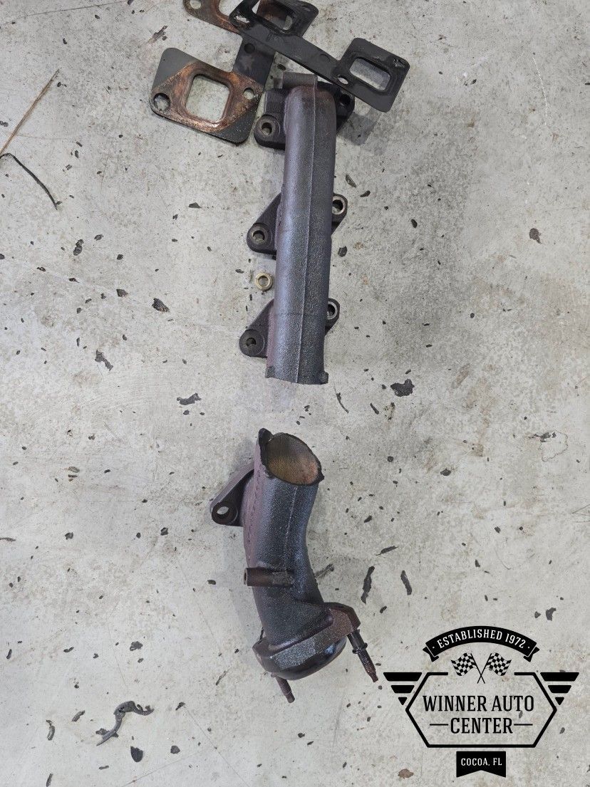 Two pieces of a rusted exhaust manifold with mounting brackets and gaskets | Winner Auto Center