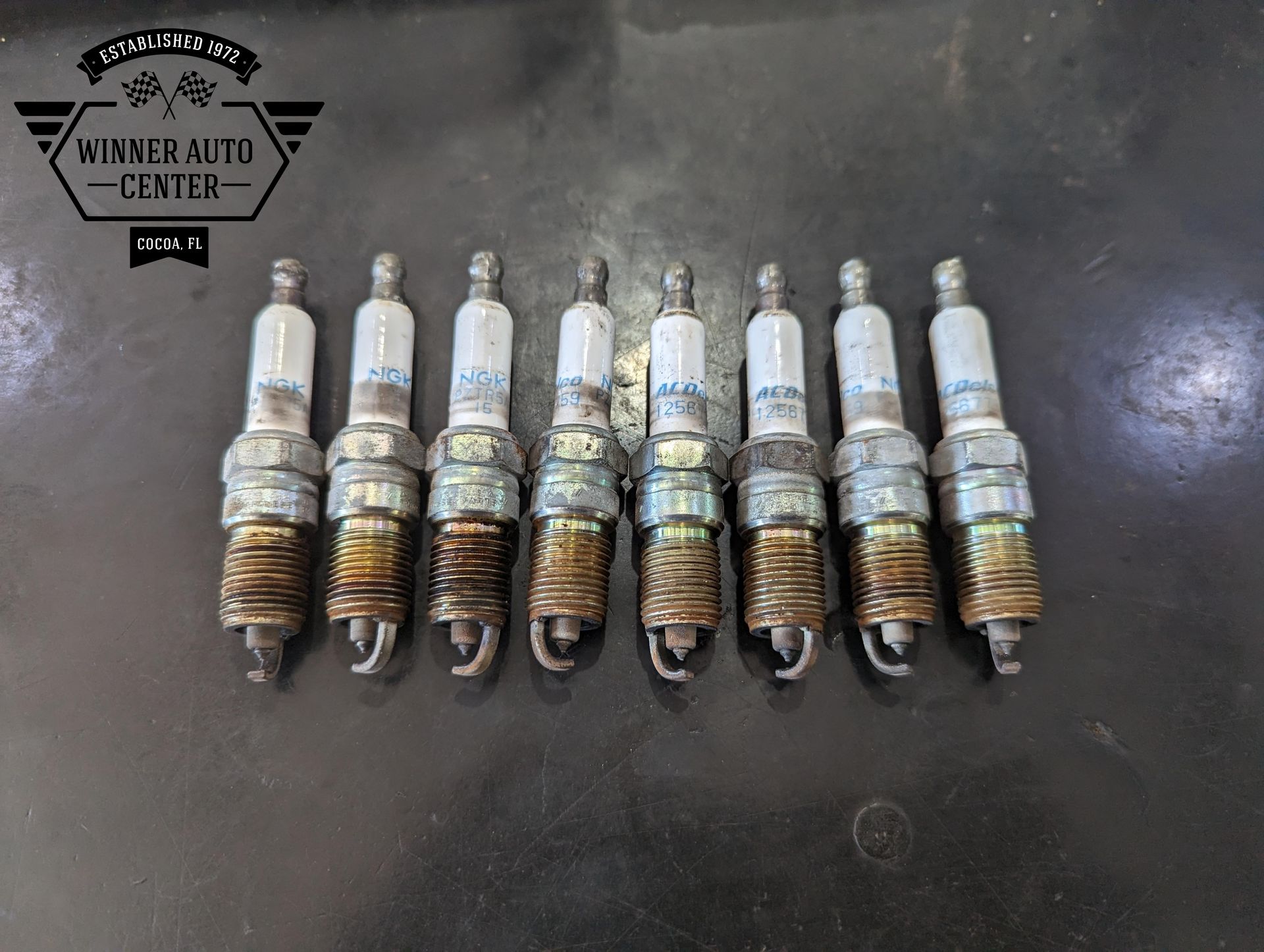 Nine used spark plugs laid out on a dark surface | Winner Auto Center