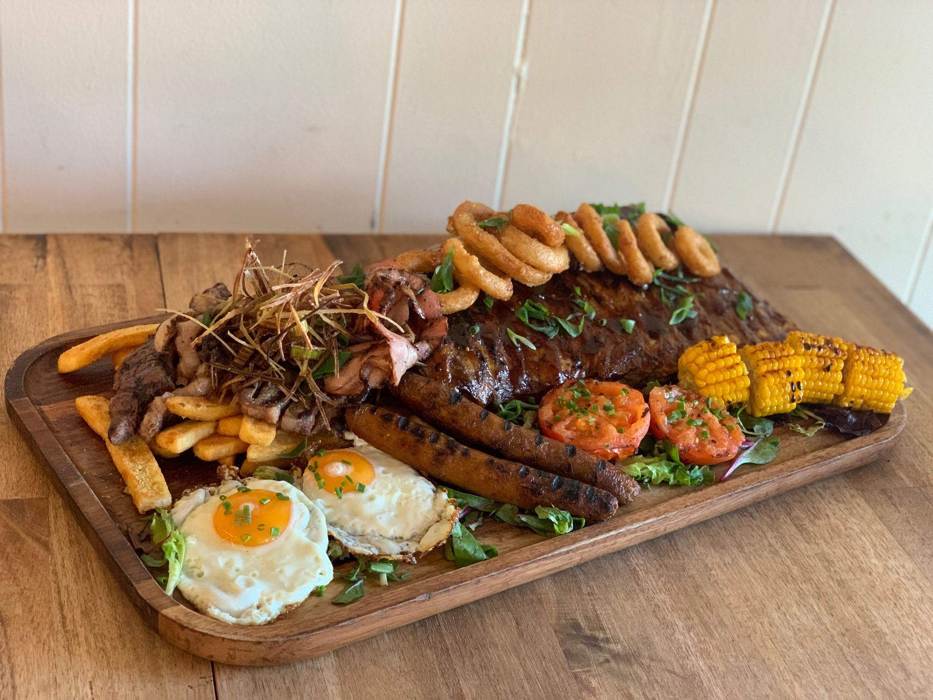A wooden cutting board topped with meat , eggs , corn on the cob and onion rings.