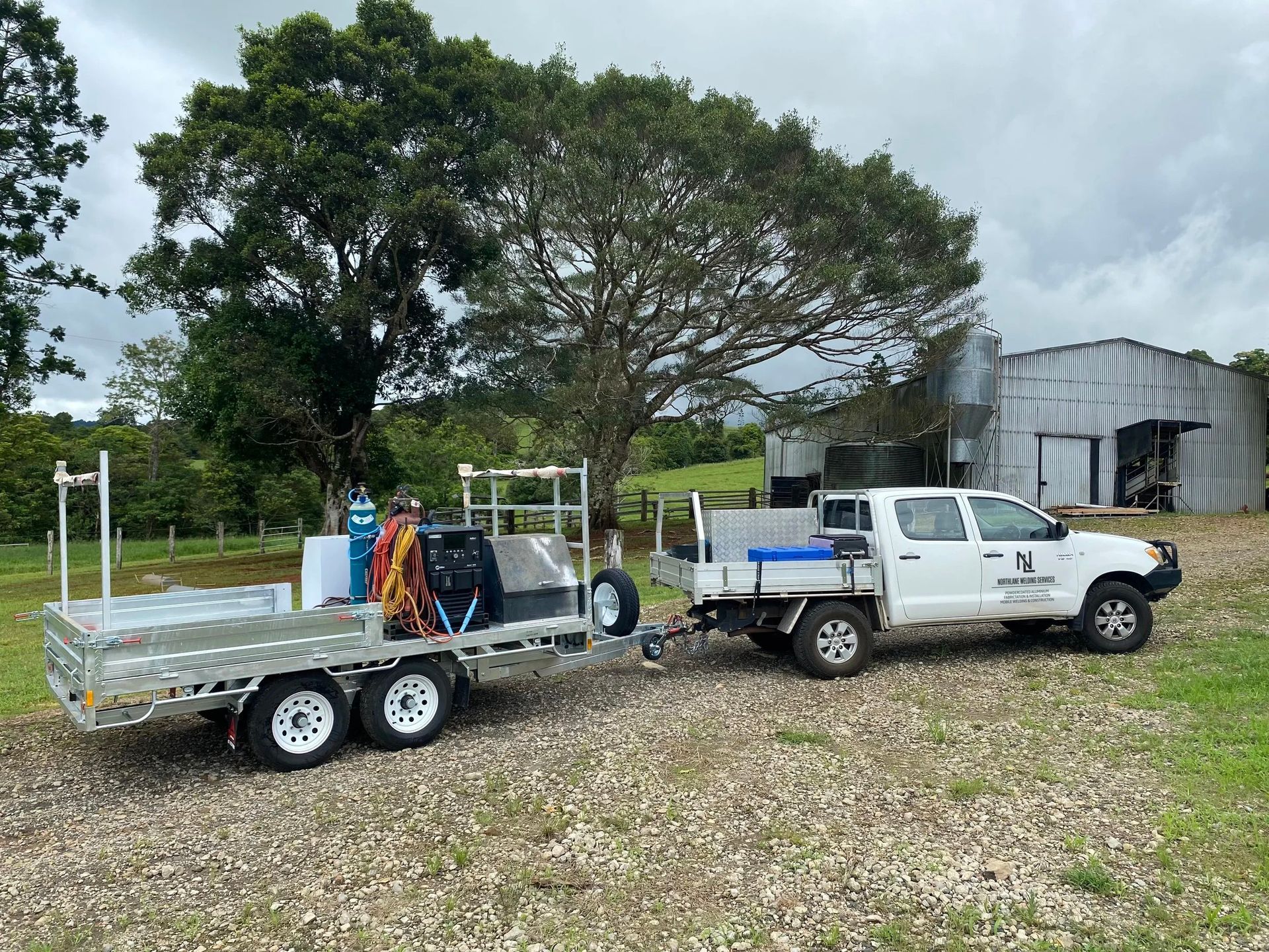 A white utility truck towing a flatbed trailer with equipment parked on a gravel area near a tree and farm building.