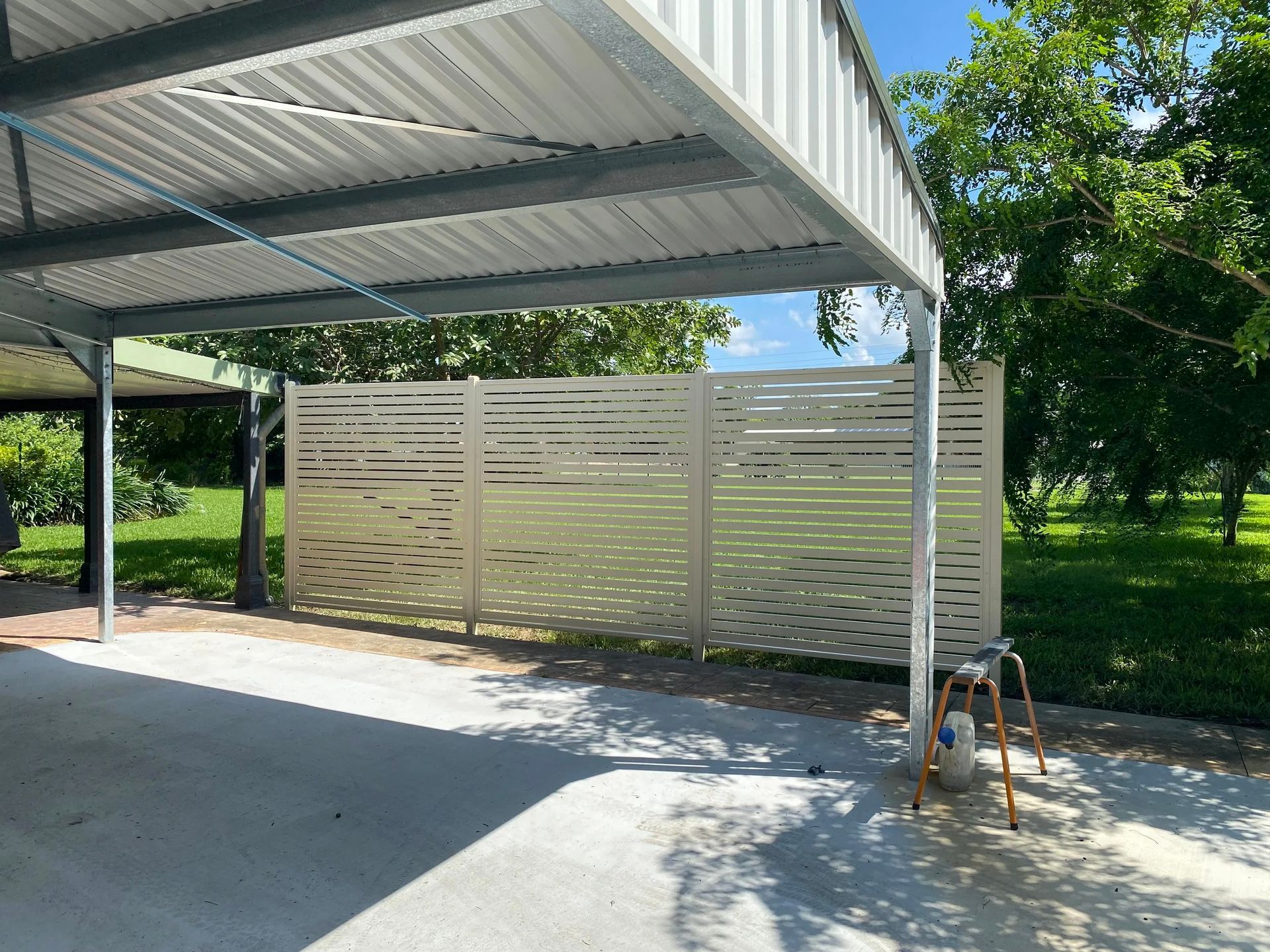 A wide, light-colored horizontal slat privacy screen stands under a metal carport canopy on a concrete slab.