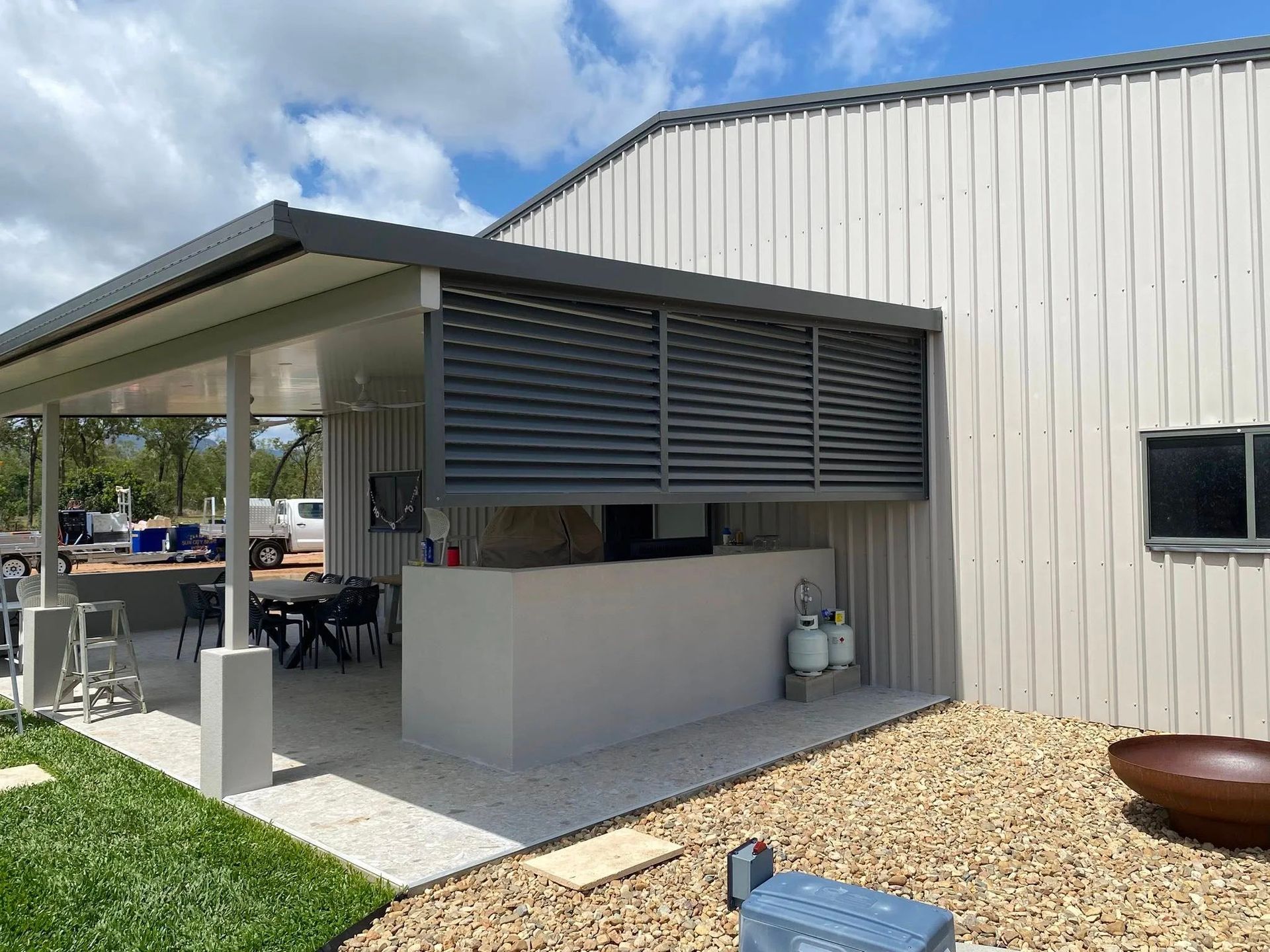 An outdoor patio area with a white bar counter, dark grey privacy louvers, and a table set under a covered metal roof.