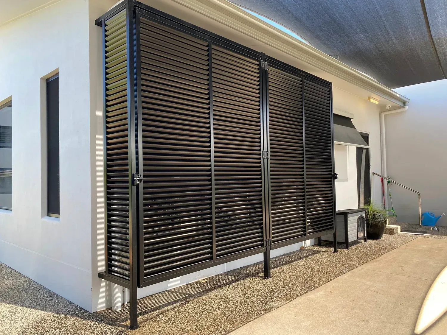 Black aluminum louvered screen wall attached to the exterior of a white house, shielding an outdoor HVAC unit.