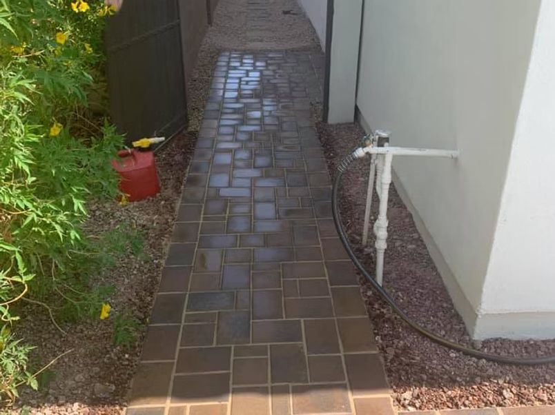 Brick pathway between a green bush and a white wall, with gravel edges and utility pipes.
