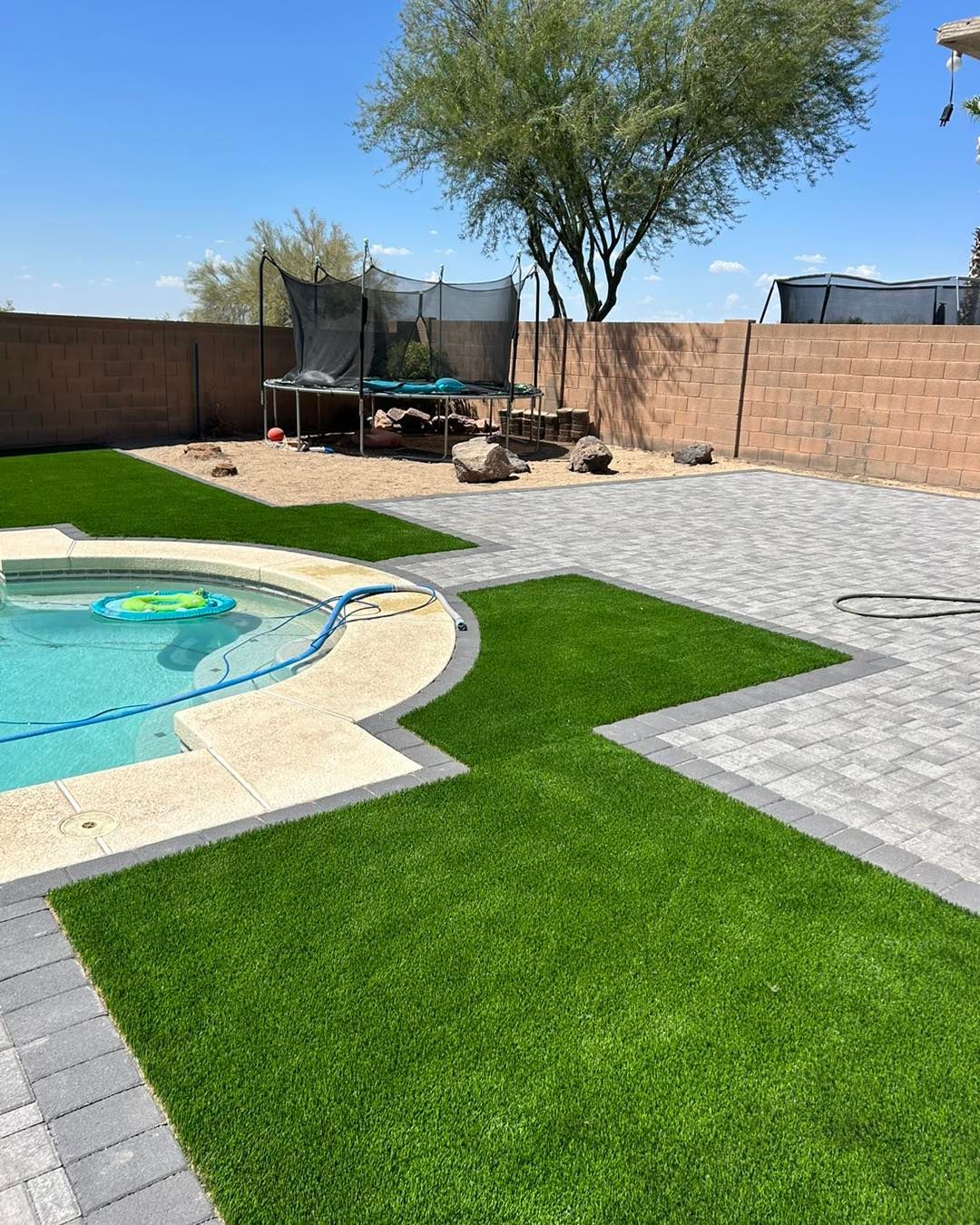 Backyard with pool, artificial turf, and paved patio; trampoline and tree in the background.