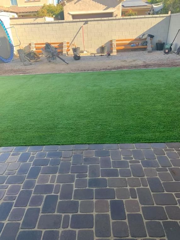 Brick patio leads to green artificial turf lawn; background wall with landscaping.