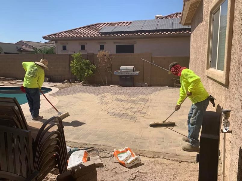 Two workers in neon shirts and hats sweeping a concrete patio near a pool, a house, and a grill.