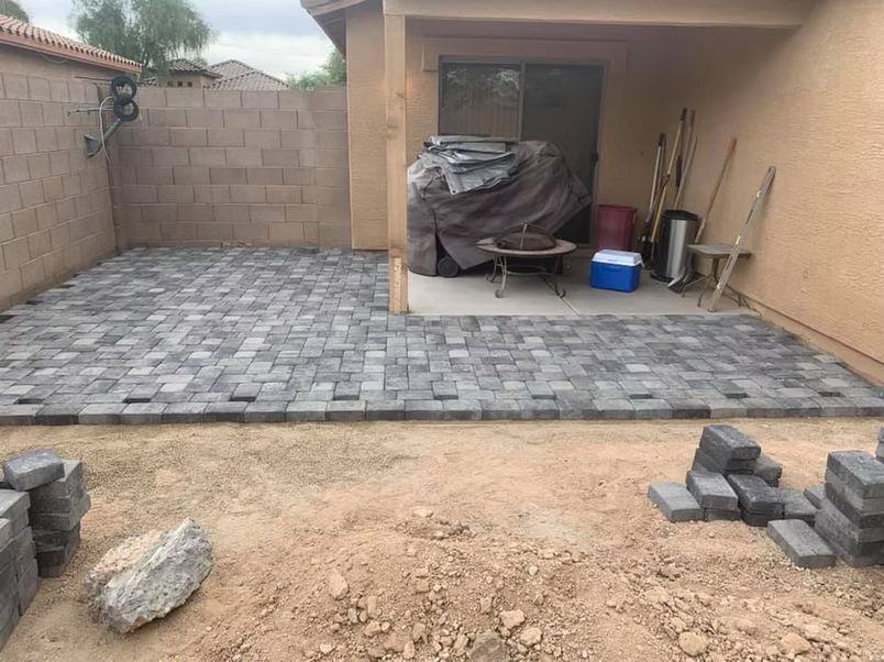 Patio pavers being installed in a backyard. Unfinished section with dirt, finished section of gray pavers.