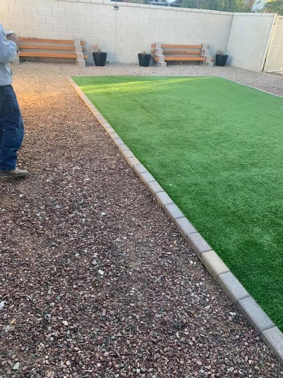 Gravel path next to artificial turf yard with benches and potted plants against a wall.