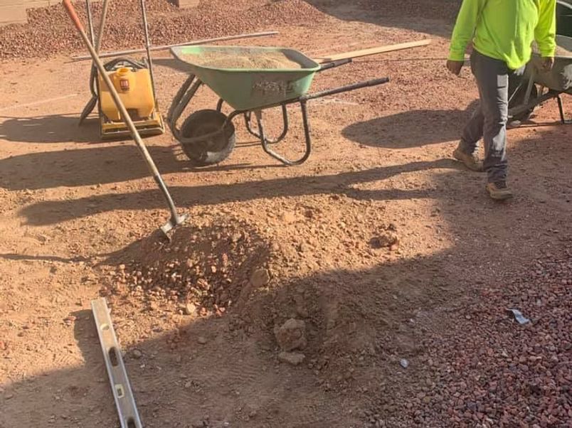 Construction worker near a wheelbarrow, tamp, and dirt pile on a gravel surface.